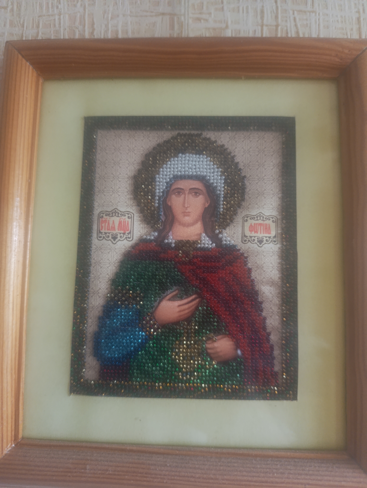 A framed, beaded religious icon, depicting a female saint, hangs on a textured, light-colored wallpapered wall in the city of Vasylkiv, Ukraine. The central image features a bust-length rendering of a saint with a printed face and visible right hand, surrounded by a halo of white and gold beads. Her attire, comprising a red mantle draped over a green and blue garment, is meticulously crafted from small, shimmering beads. Inscriptions in Cyrillic, "СТАА МЦА" (Holy Martyr) and "ФΨТIНА" (Photina or Svetlana), appear on either side of her head. The beaded artwork is framed with a dark beaded border, mounted within a light yellow-green matte, and enclosed by a light-toned, wooden frame with mitered corners.