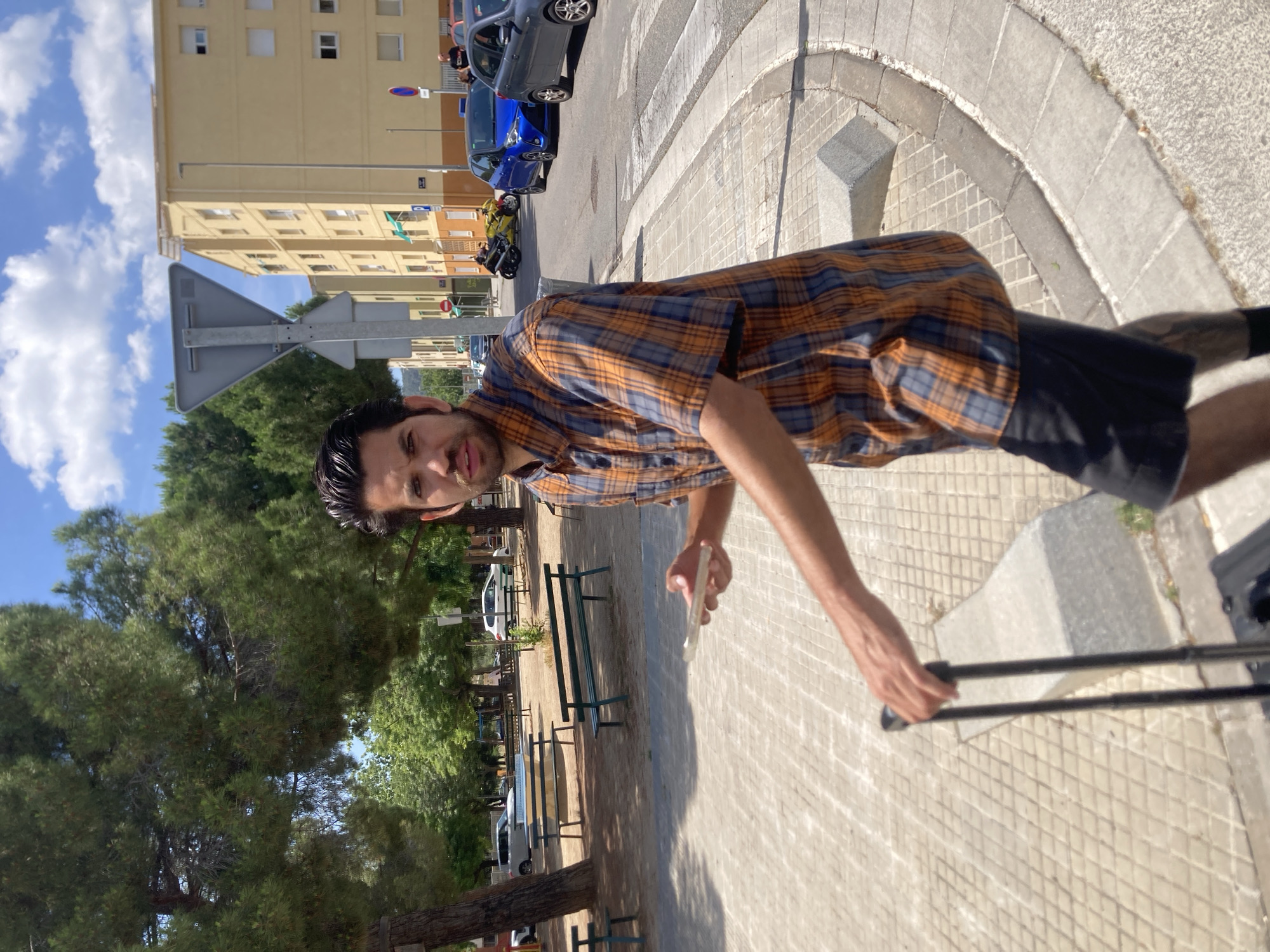 A young man with dark hair and a beard stands on a paved sidewalk in Terrassa, Spain, holding a cell phone and a suitcase. He is wearing a plaid shirt with orange, blue, and white squares, and black shorts. The man is looking at the camera with a neutral expression. In the background, there are trees, buildings, parked cars, and a street with a crosswalk. The sky is blue with scattered clouds. The sun is shining, casting shadows on the ground.