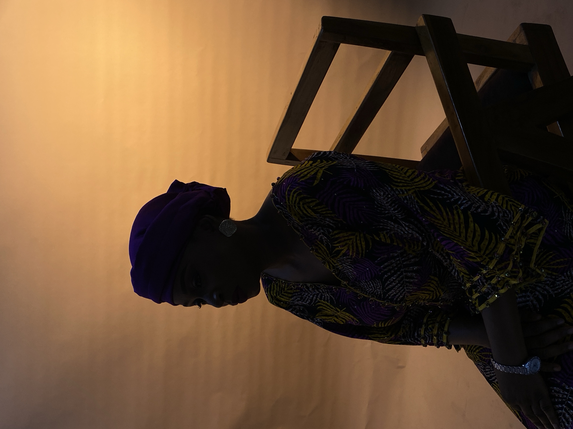 A woman wearing a purple head wrap and a patterned dress is positioned in front of an orange backdrop, with a wooden chair frame partially obscuring the upper right of the frame. She is wearing a watch on her left wrist and an earring. The scene is underexposed. The location is Gura Top, Nigeria.