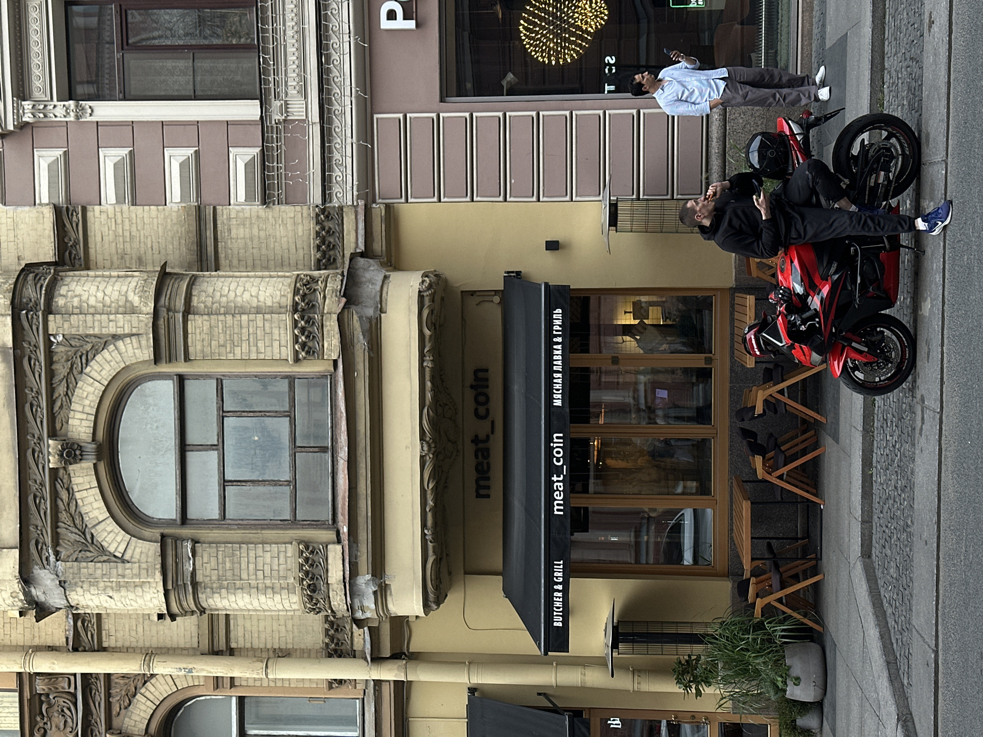 This image depicts a street scene in Saint Petersburg, Russia, in front of a building with a light yellow facade and intricate architectural details. A black awning displays the text "meat_coin" in white lettering, along with "BUTCHER & GRILL" and Cyrillic script. To the right of the awning, a large window reveals the interior of a establishment.
In the foreground, a red motorcycle is parked on the cobblestone street. A man wearing a black hoodie and dark pants is seated on the motorcycle, eating and holding a phone. To the right, another man in a light blue striped shirt and grey pants stands looking at his phone. Outdoor seating with wooden tables and chairs is visible on the sidewalk in front of the establishment. The background shows the upper floors of the building with arched windows and decorative elements. A tall potted plant with green foliage is positioned near the entrance.