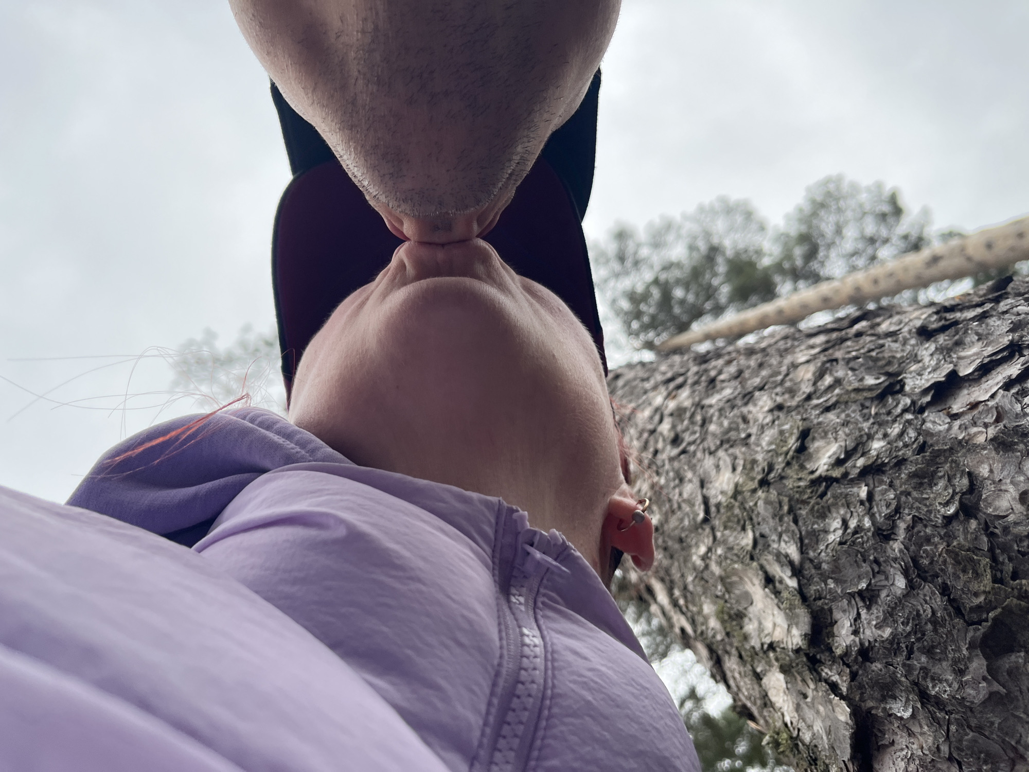 A low-angle shot shows a couple kissing outdoors against a cloudy sky. The person in the foreground wears a lilac jacket and has a gold earring in their left ear. The person behind them wears a black baseball cap. A tree trunk with rough bark is visible to the right of the couple. The location is Chaikovsky, Russia.