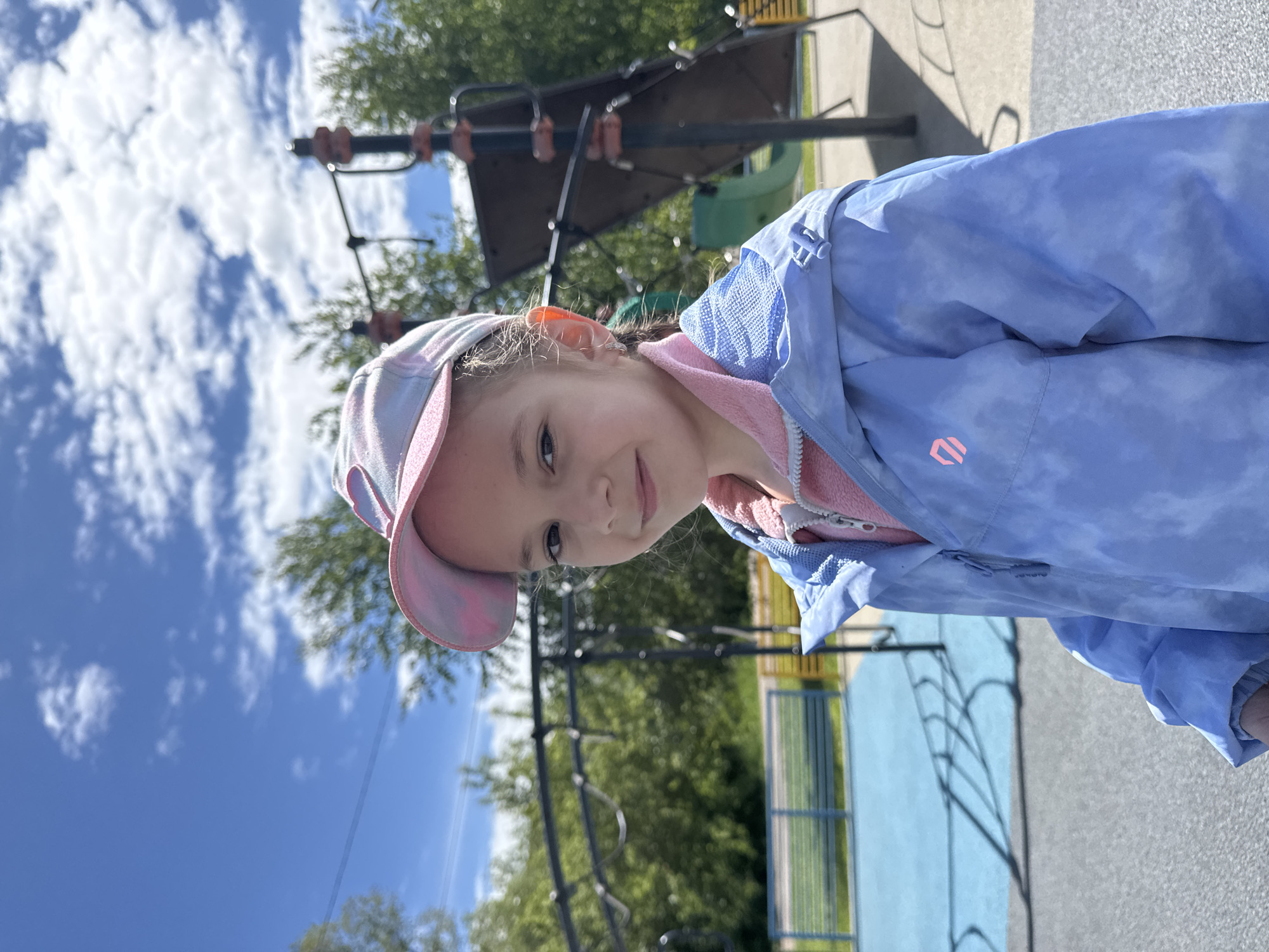 A young girl with light brown hair stands at a playground in Shevlyakovo, Russia. She wears a light blue and pink patterned cap and a light blue jacket over a pink shirt. Behind her is a playground structure made of metal bars and a green slide. A blue painted surface is adjacent to a gray surface, separated by a green fence. The sky is blue with scattered white clouds. Trees are visible in the background.