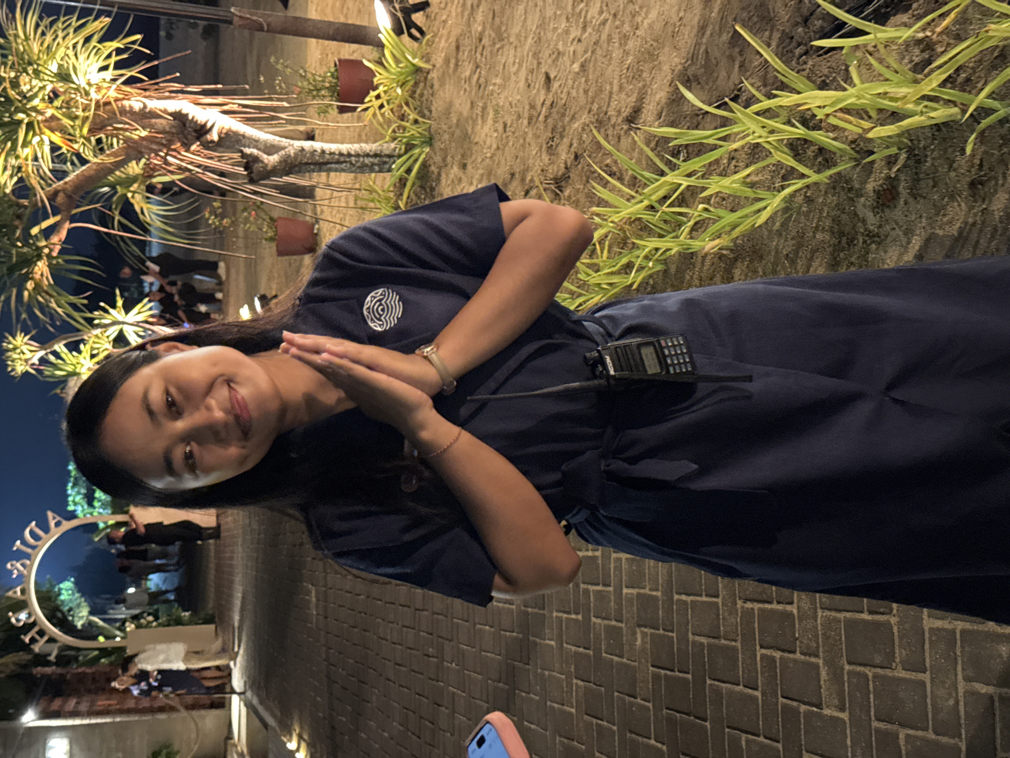 A young adult female stands centrally, facing forward and smiling, with dark hair. She wears a dark blue short-sleeved uniform dress featuring a tie-waist and a white circular logo with a wave motif on the left chest. A black ICOM walkie-talkie is clipped to her left side. Her hands are clasped together at chest level in a traditional greeting gesture, with a light-colored bracelet visible on her right wrist.
The environment is an outdoor setting at night, illuminated by artificial warm lighting. To the left of the subject is a dark brick or stone paved pathway. A light pink object, possibly a phone case, is partially visible on this path in the lower left foreground. In the background, an illuminated arched gateway with the mirrored text "ADIA RHYA" (or similar) is visible on the left, leading to an area occupied by several blurred human figures. To the right of the subject, the ground consists of sandy soil, featuring rows of light green, spiky plants and several potted plants. Multiple tropical trees with distinctive spiky foliage are present, illuminated by spotlights from below. The scene is located in Nusa Dua, Indonesia.