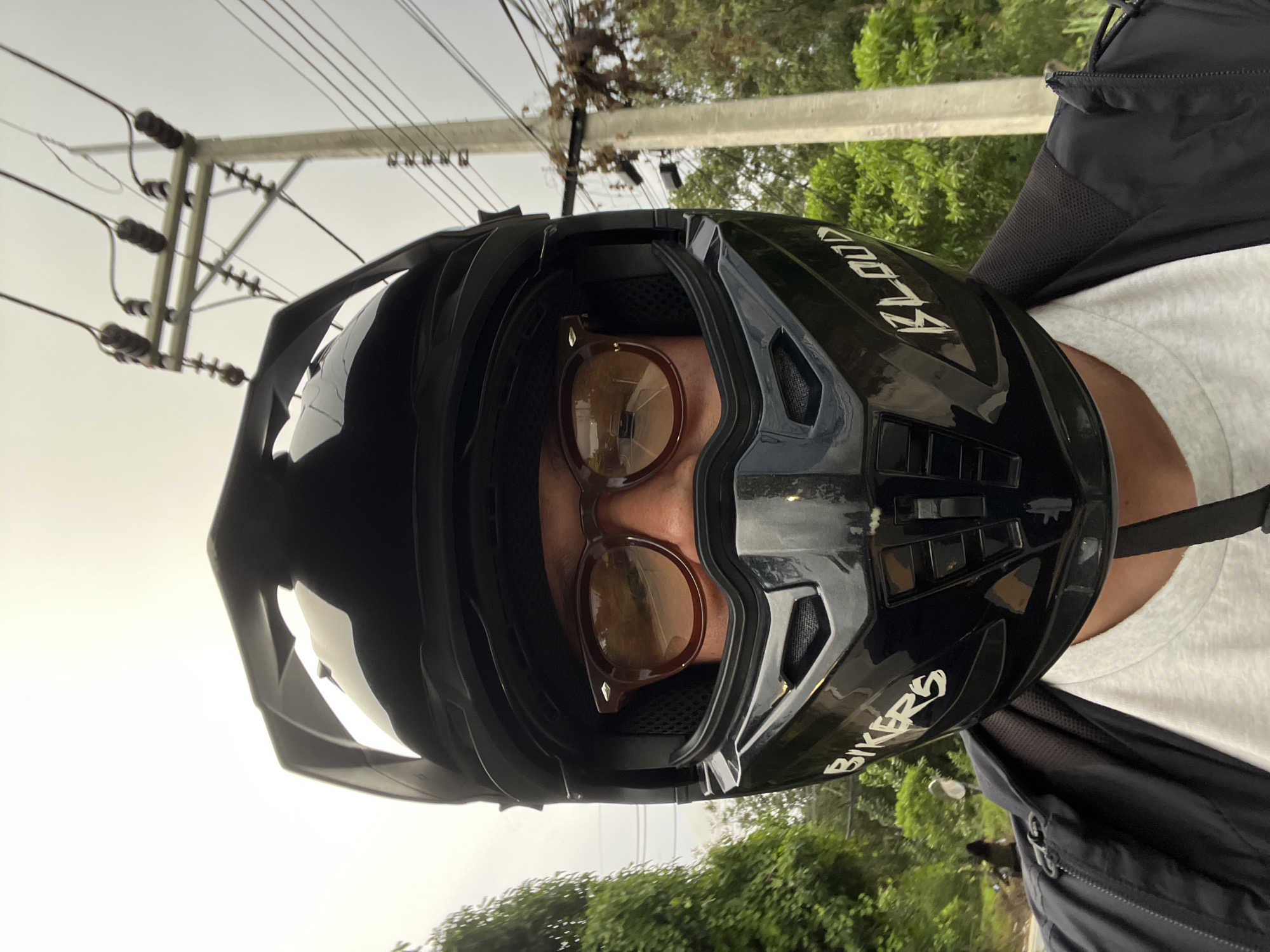 A medium shot shows a person wearing a black motorcycle helmet with a visor and brown-tinted glasses. The helmet has the words "BIKER'S" and "BLOOD" printed on it in white. The person is wearing a white t-shirt and a black jacket. In the background, there are trees, a utility pole with power lines, and a road with a vehicle in the distance. The sky is overcast. The image appears to have been taken outdoors in a semi-rural or suburban area.