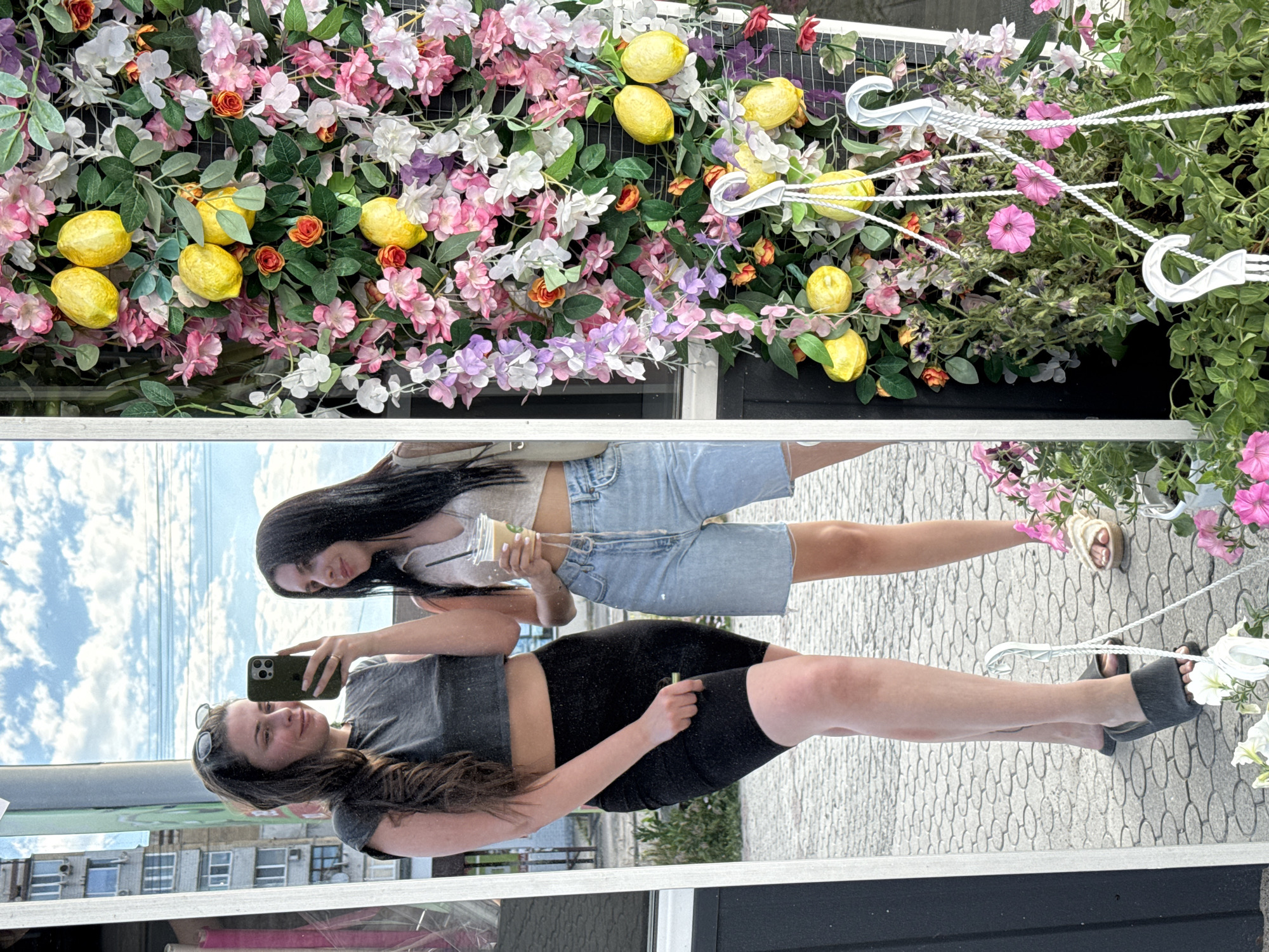 Two young women are captured in a mirror selfie, standing in front of a wall adorned with artificial lemons and flowers. The woman on the left, with long, dark hair and sunglasses perched on her head, wears a grey crop top and black bike shorts. She holds a dark-colored smartphone, taking the photo. The woman on the right, also with long dark hair, is dressed in a light-colored crop top and denim shorts. She holds a takeaway cup with a straw. They are both wearing casual footwear. The background visible in the reflection shows a city street with buildings and a cloudy sky. To the right of the women, the floral and lemon display is prominent, with various colors of flowers and yellow fruit arranged on a black mesh backing. Hanging planters with more pink flowers are also visible. The ground beneath them is paved with a cobblestone-like pattern.
