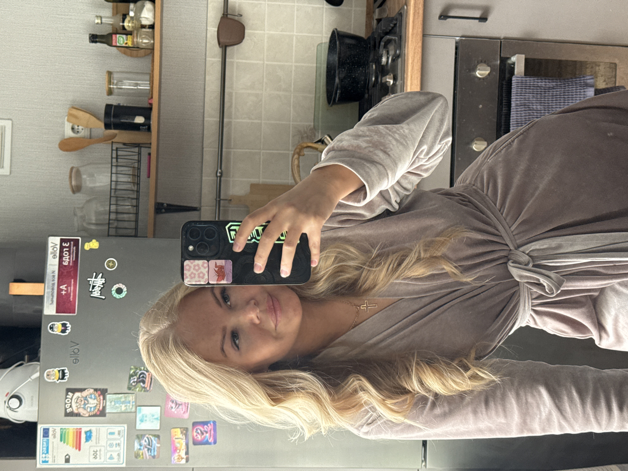 A young woman with long, wavy blonde hair is taking a selfie in what appears to be a kitchen. She is wearing a light brown, long-sleeved dress or robe with a tied waist. She is holding a smartphone with a black case. The background reveals a kitchen setting with cabinets, appliances, and cooking utensils. A refrigerator is visible on the left, adorned with various magnets and stickers. On the right, there's a stove with a black pot and other kitchen items. The overall lighting is bright and appears to be natural.
