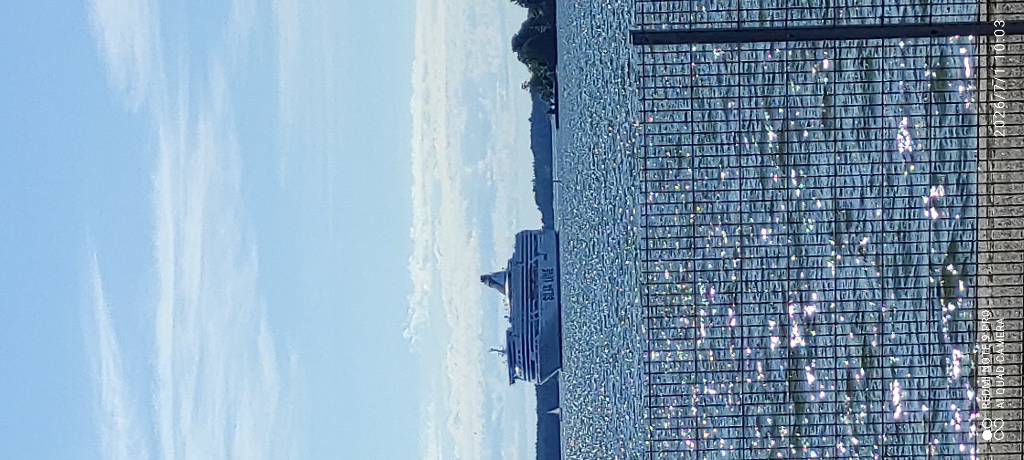 The image shows a ferryboat named "SILA LINE" sailing on a body of water, likely the sea or a large lake. The ferry is white and appears to be quite large, with multiple decks visible. It is seen from the side, with its bow pointing towards the left of the frame. The water is choppy, reflecting the sunlight and creating a shimmering effect.
In the background, a tree-covered coastline is visible, suggesting the ferry is near land. The sky is blue with scattered white clouds, indicating a bright and clear day. A fence or railing with a grid pattern is in the foreground on the right side of the image, partially obscuring the view of the water and the ferry. This fence has a texture that catches the light, adding to the overall visual effect.
Text is visible in the bottom right corner of the image, which reads "REDMI NOTE 9 PRO AI QUAD CAMERA" and a timestamp "2026/7/1 10:03". The timestamp suggests the image was taken in the morning on July 1st, 2026. The scene evokes a sense of travel and pleasant weather.