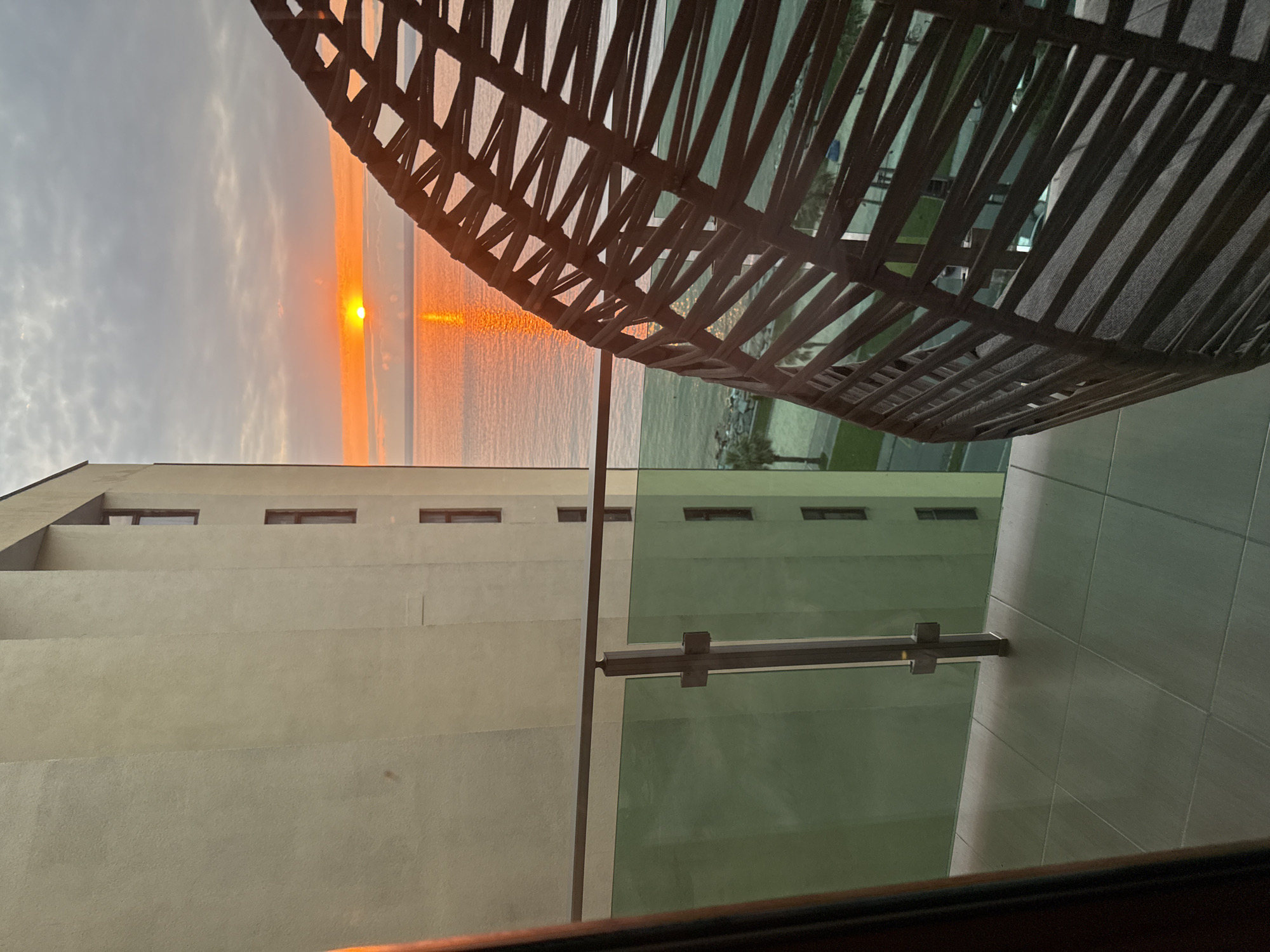 The image depicts a view from a balcony overlooking the Black Sea at sunset in Batumi, Georgia. A modern building with beige exterior walls and dark-framed windows occupies the left side of the frame. A glass balcony railing with metal supports is visible in the foreground, with the tiled floor of the balcony below. In the center of the frame, the sun is setting over the calm sea, casting a warm orange glow across the water and the sky. A large, woven, egg-shaped chair with a dark frame partially obstructs the view on the right side of the image. In the distance, a green expanse suggests a park or promenade with palm trees lining a road.