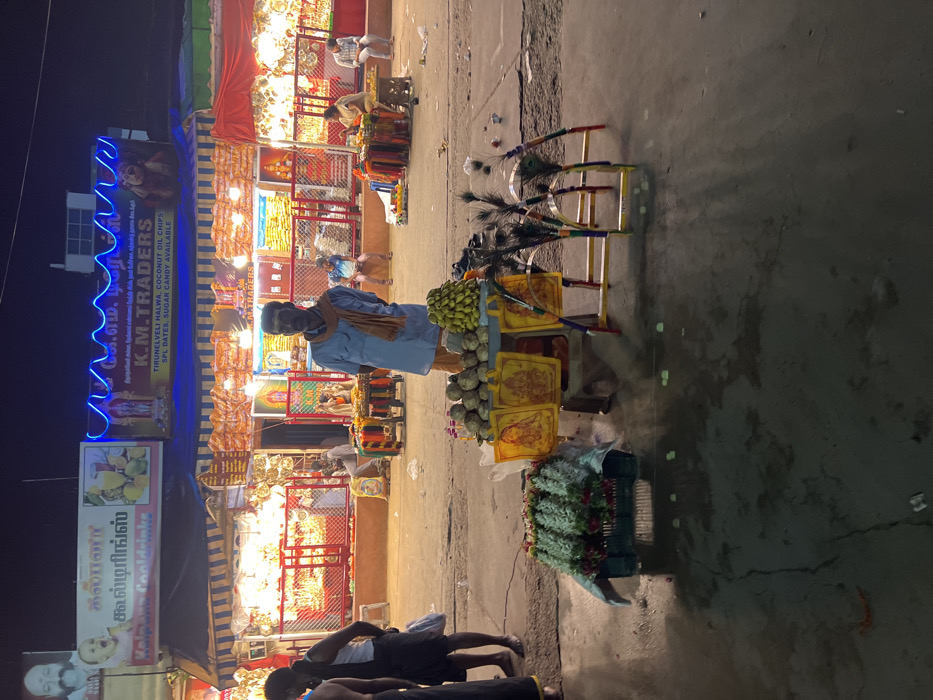 The image captures a vibrant night street scene, likely in a market area in Palani, India. The sky is dark, indicating nighttime, and the environment is brightly lit by numerous artificial lights from shops and vendor stalls, along with decorative blue string lights.
In the foreground, a man stands behind a mobile cart, appearing to be a vendor. He wears a blue shirt, a scarf, and a face mask. His cart displays an array of goods including bunches of green bananas, piles of what look like limes or small fruits, and several framed images, possibly devotional pictures of Hindu deities. Tall sticks adorned with peacock feathers are also arranged on his cart. On the ground next to his cart, a basket holds what appear to be garlands or flowers, commonly used for religious offerings.
Behind the vendor, a row of shops or permanent stalls lines the street. These establishments are brightly illuminated, showcasing their wares which appear to include packaged goods, decorative items, and possibly religious paraphernalia. Several other people are visible near and inside these shops, suggesting a bustling commercial atmosphere.
Visible text on the shop signs includes "K.M. TRADERS," with a more detailed sign listing products like "TIRUNELVELI HALWA, COCONUT OIL CHIPS, SPL DATES, SUGAR CANDY AVAILABLE." Another sign reads "கல்பனா கூல்டிரிங்ஸ்" (Kalpana Cooldrinks). The combination of devotional items, traditional South Indian snacks, and drinks points to a lively market street, characteristic of towns, especially those with significant temples like Palani. The scene conveys a sense of active nocturnal commerce and community life.