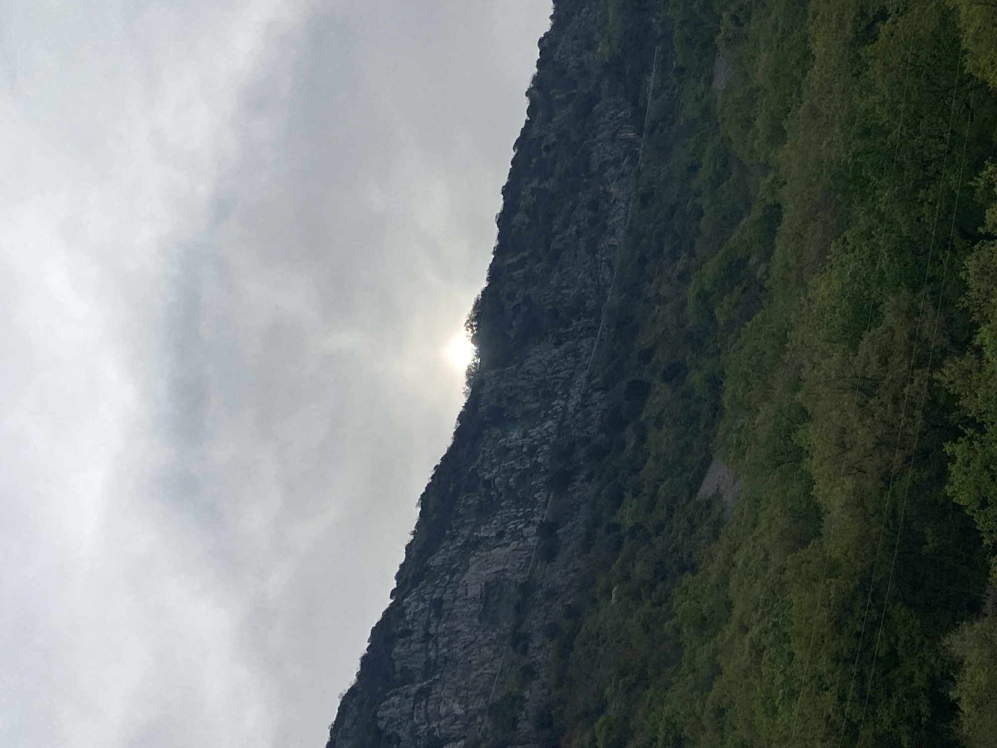 The image shows a mountainside on a cloudy day. The sun is partially obscured by the mountain. The mountain is covered in trees and rocky terrain. The image could potentially be in the city of Budva, Montenegro.