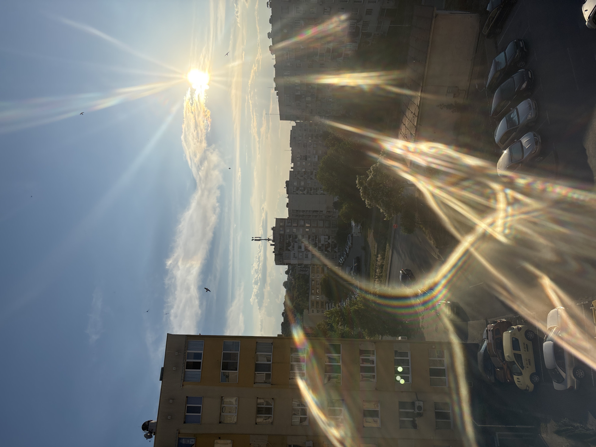 The image captures a view of urban buildings and a sky with the sun shining brightly. In the foreground, on the left, is a tall, light-colored apartment building with multiple windows. Below and to the right, several parked cars are visible, including a yellow taxi. Beyond these, a street with a few cars is seen, leading towards a series of taller, gray apartment buildings. The sky is blue with scattered white clouds, and the sun is positioned in the upper right quadrant, casting strong rays and lens flare across the scene. Several birds are visible flying in the sky. The overall impression is that of a late afternoon or early evening urban landscape. This scene is located in Dupnitsa, Bulgaria.