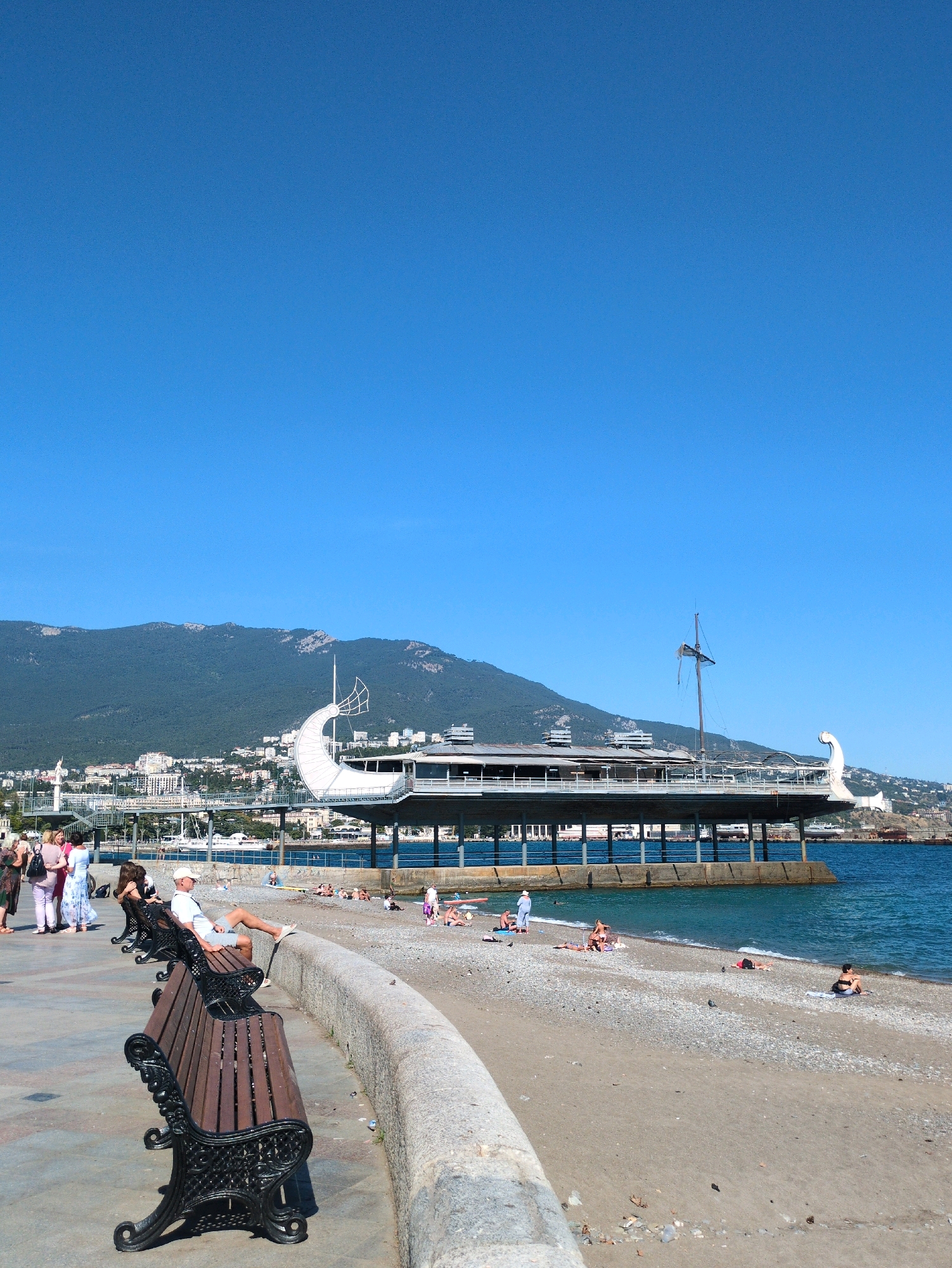 The image depicts a seaside promenade with a beach and a building structure extending over the water. In the foreground, two park benches with dark wood slats and ornate black metal frames are positioned on a paved walkway. A man wearing a white hat, white shirt, and grey shorts is seated on the bench closest to the camera, with his legs extended and resting on a curved stone wall that separates the walkway from the beach. To his right, on the beach, several individuals are sunbathing on the sand and pebbles. The beach is bordered by the turquoise water of the sea. In the mid-ground, a long, elevated building with a modern design, featuring large windows and a curved white architectural element resembling a sail or a crescent moon, extends over the water. A flag on a mast is visible atop the structure. In the background, a green, tree-covered mountain rises behind a city with buildings of various sizes and colors. The sky is clear and blue. A few pigeons are present on the beach.