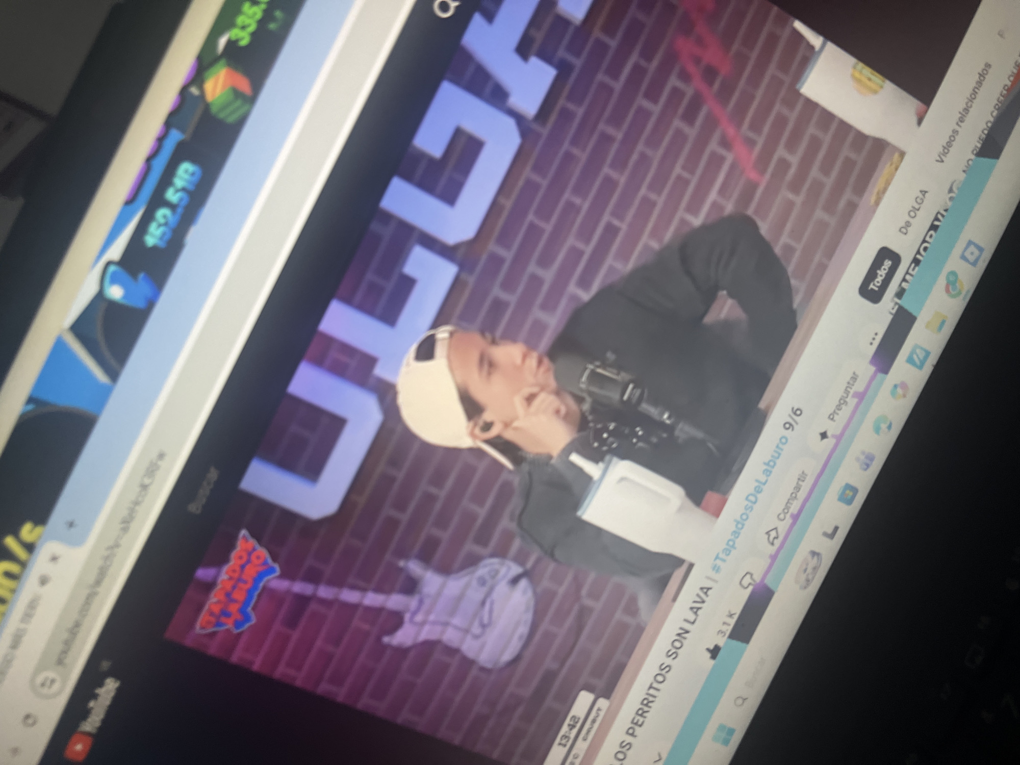 The image shows a person wearing a baseball cap and headphones, speaking into a microphone. They are in front of a brick wall with large white letters that spell "ULUGAR". A guitar is visible on the wall behind them. The person appears to be on a live stream or recording a video, as indicated by the YouTube interface visible on the screen. The timestamp on the screen reads "13:42". The overall environment appears to be a studio or a room set up for broadcasting. The image does not contain any information or visual cues that would place the location in Santa Ana, Venezuela.
