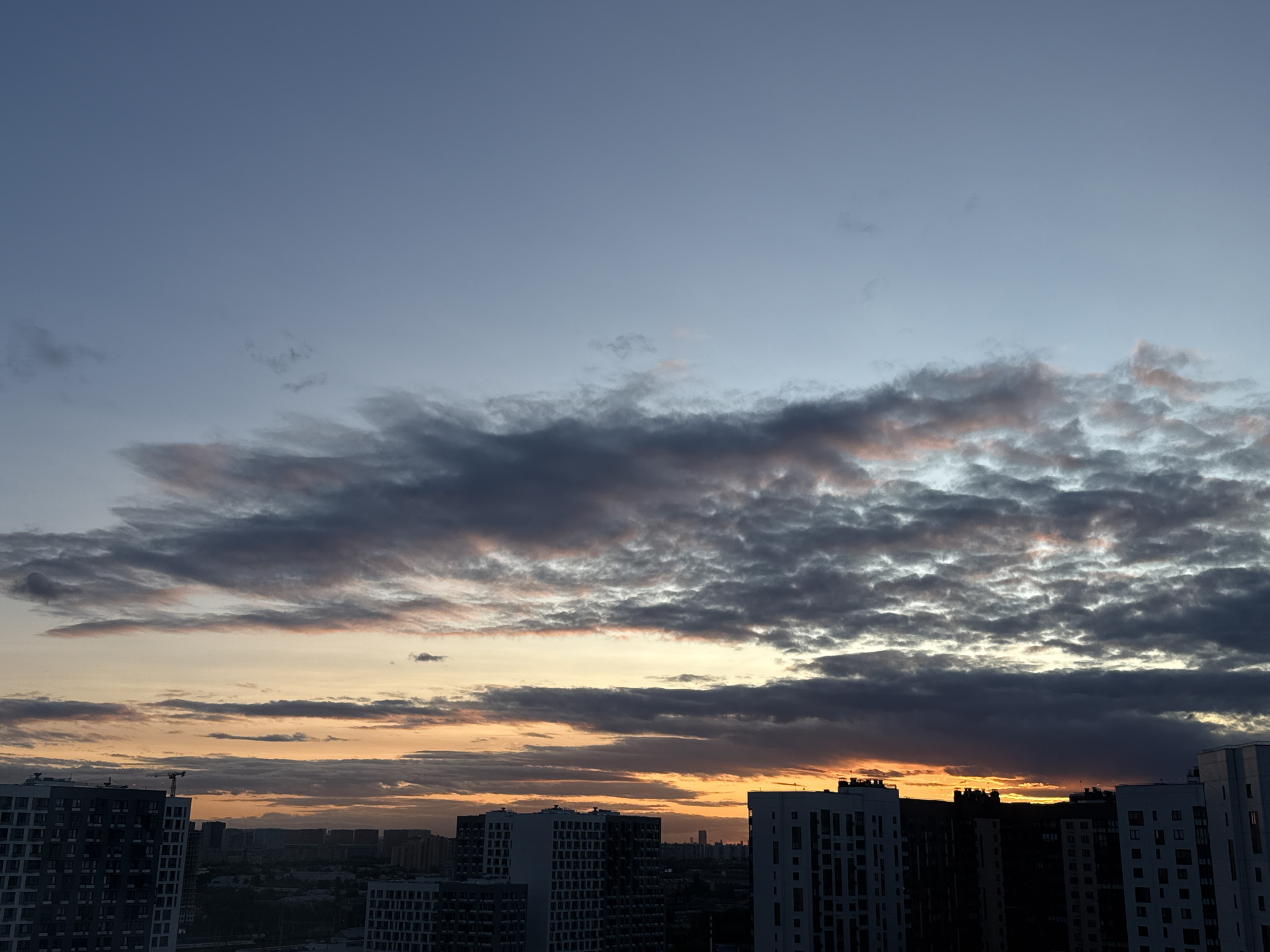 The image shows a panoramic view of the city of Lyubertsy, Russia, during a sunset. The sky is a gradient of colors, transitioning from a light blue at the top to hues of orange and yellow near the horizon, with dark clouds scattered across. Silhouetted against the colorful sky are several tall apartment buildings, characteristic of modern urban development. A construction crane is visible on top of one of the buildings on the left side of the frame, indicating ongoing construction. The cityscape extends into the distance, with more buildings and a hazy horizon. The overall scene depicts a typical urban environment at dusk, with the dramatic colors of the sunset providing a striking backdrop to the residential architecture.