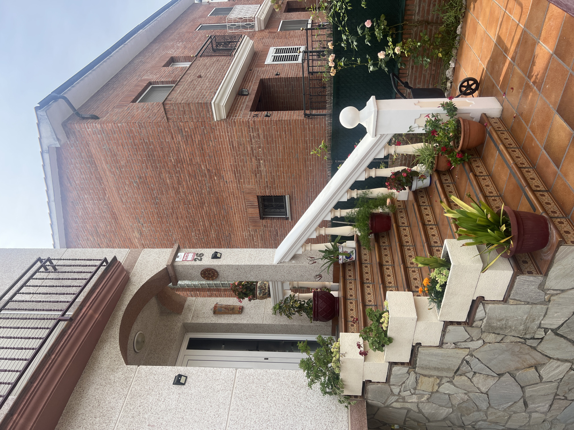 This media file captures a residential scene in Mollet del Vallès, Spain, primarily focused on the entrance of a building and a larger brick building behind it. The image is taken during the daytime under clear skies.
In the foreground, a welcoming set of stairs leads up to the entrance of a building with a light-colored, textured facade, possibly stucco or roughcast. The stairs feature risers adorned with intricate patterned tiles and terracotta-colored treads. A white balustrade with a smooth handrail and ornate spindles lines the right side of the staircase. Numerous potted plants and colorful flowers are arranged along the steps and on the landings, adding vibrant greenery to the scene. The lower portion of the stair structure and an adjacent wall are clad with irregular grey stone. A white door marks the main entrance of this building.
Behind the foreground building stands a taller, multi-story structure constructed from reddish-brown brick. This building has multiple windows with white frames, some with external shutters or grilles, and decorative white cornices and balconies with black wrought-iron railings. A black rain gutter is visible along its roofline. Adjacent to the brick building, rose bushes with light-colored blooms and other lush greenery contribute to a garden-like atmosphere. A small black wheeled cart or garden tool rests on the tiled landing near the top of the stairs on the right.
No people are present, and no specific activity is taking place; the scene depicts a static architectural and garden view. The bright natural light indicates it is daytime. On a column next to the front door, the house number "26" is clearly visible. Below it, a red and white sticker, possibly a security alarm sign, is partially legible.