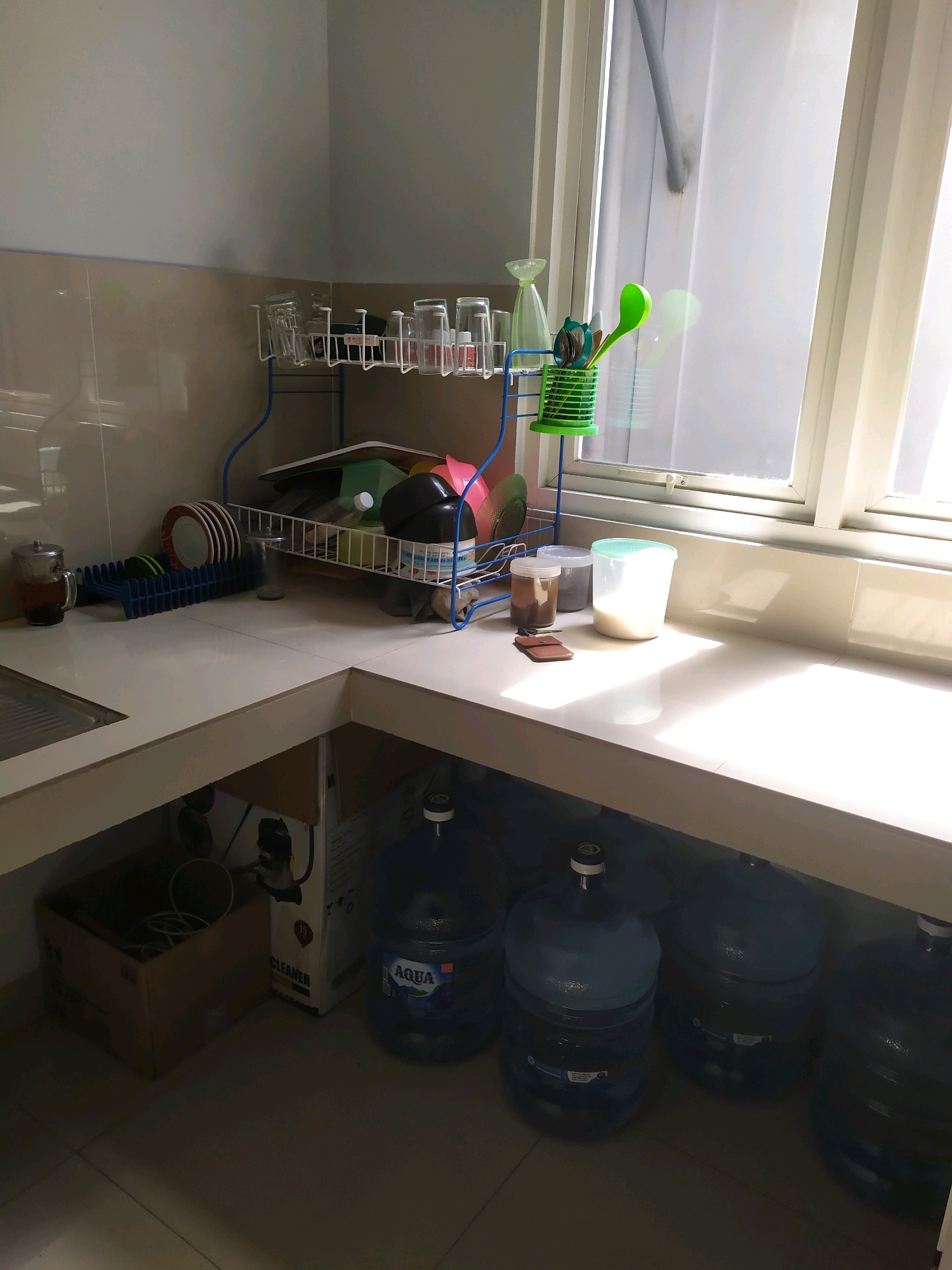 The image depicts a corner of a kitchen in what appears to be a residential setting in Wates, Indonesia. The countertop is white and L-shaped, with a sink on the left side. Above the sink, a dish rack holds several glasses and plates. To the right of the dish rack, on the countertop, are various items including jars, a container of milk, and a small brown object. A window with vertical blinds is situated on the right side of the image, allowing natural light to illuminate the scene. Below the countertop, several large blue water jugs are visible. To the left, beneath the counter, a cardboard box containing some items is present. The overall impression is of a functional, albeit somewhat cluttered, kitchen space. There are no people visible in the image.
