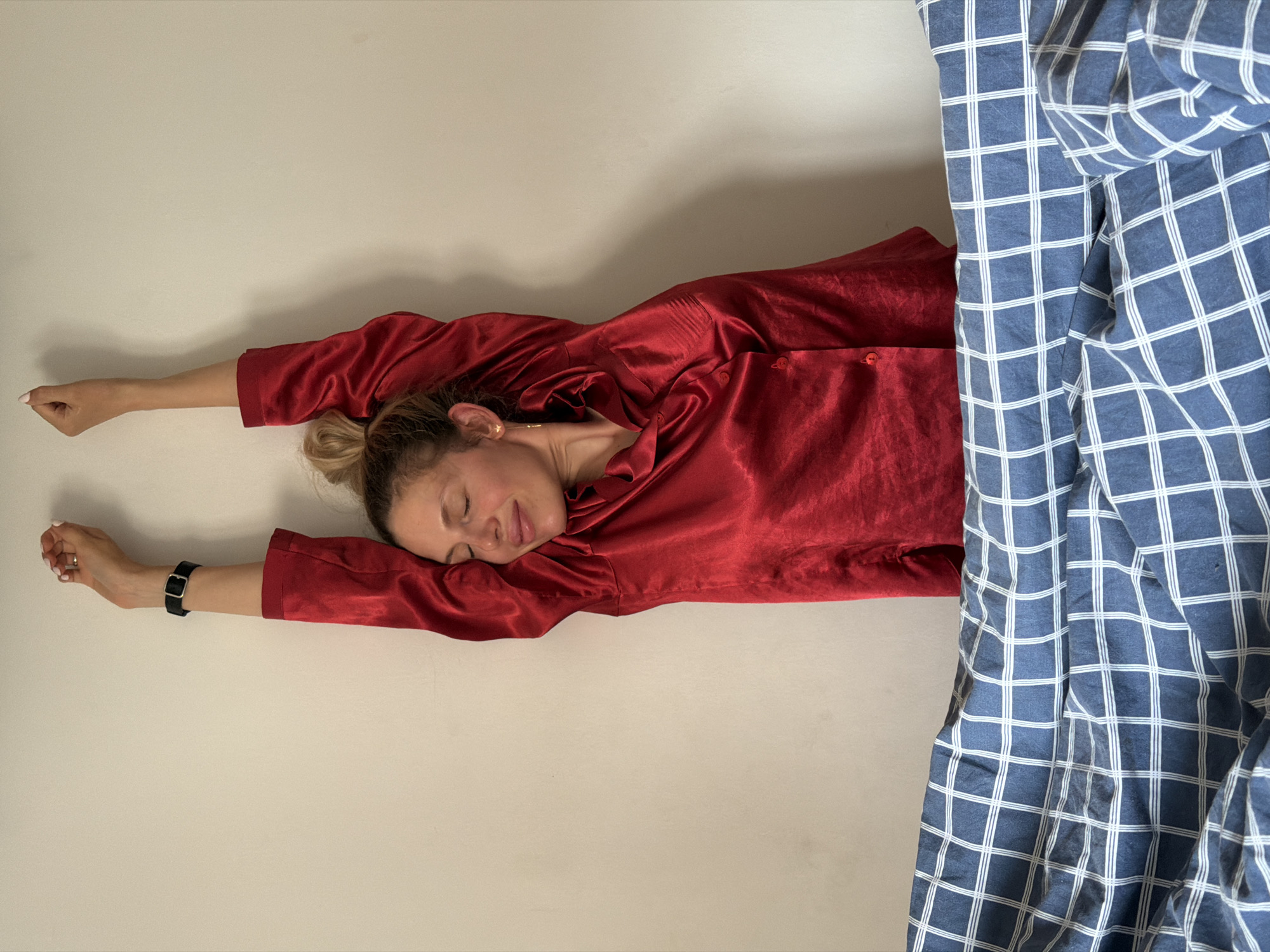 A Caucasian woman in a red pajama set lies flat on her back on a white surface that may be a bed, stretching her arms upwards. She appears to be sleeping with her eyes closed. A blue and white checkered blanket or sheet is partially covering her on the right side of the image. A black watch is visible on her left wrist. The scene is indoors, likely a bedroom. The location in Budva, Montenegro cannot be determined from this image.