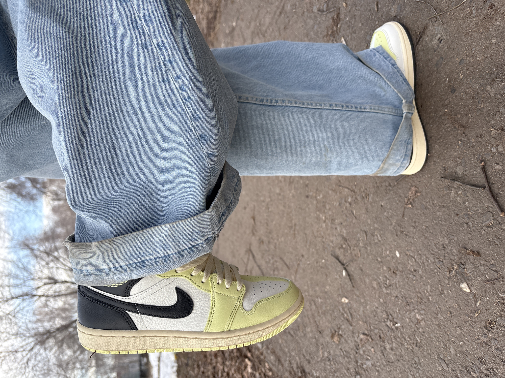 The image shows a person wearing light blue jeans and sneakers, standing on what appears to be a paved surface or possibly a dirt path. The sneakers are predominantly white and light green, with a black accent mark. The jeans are rolled up at the cuffs. The image is taken at an angle, showing only the person's legs and feet. The environment is muted, suggesting an outdoor setting. Without further details, definitively locating it in Ulyanovsk, Russia is not possible from this single image.