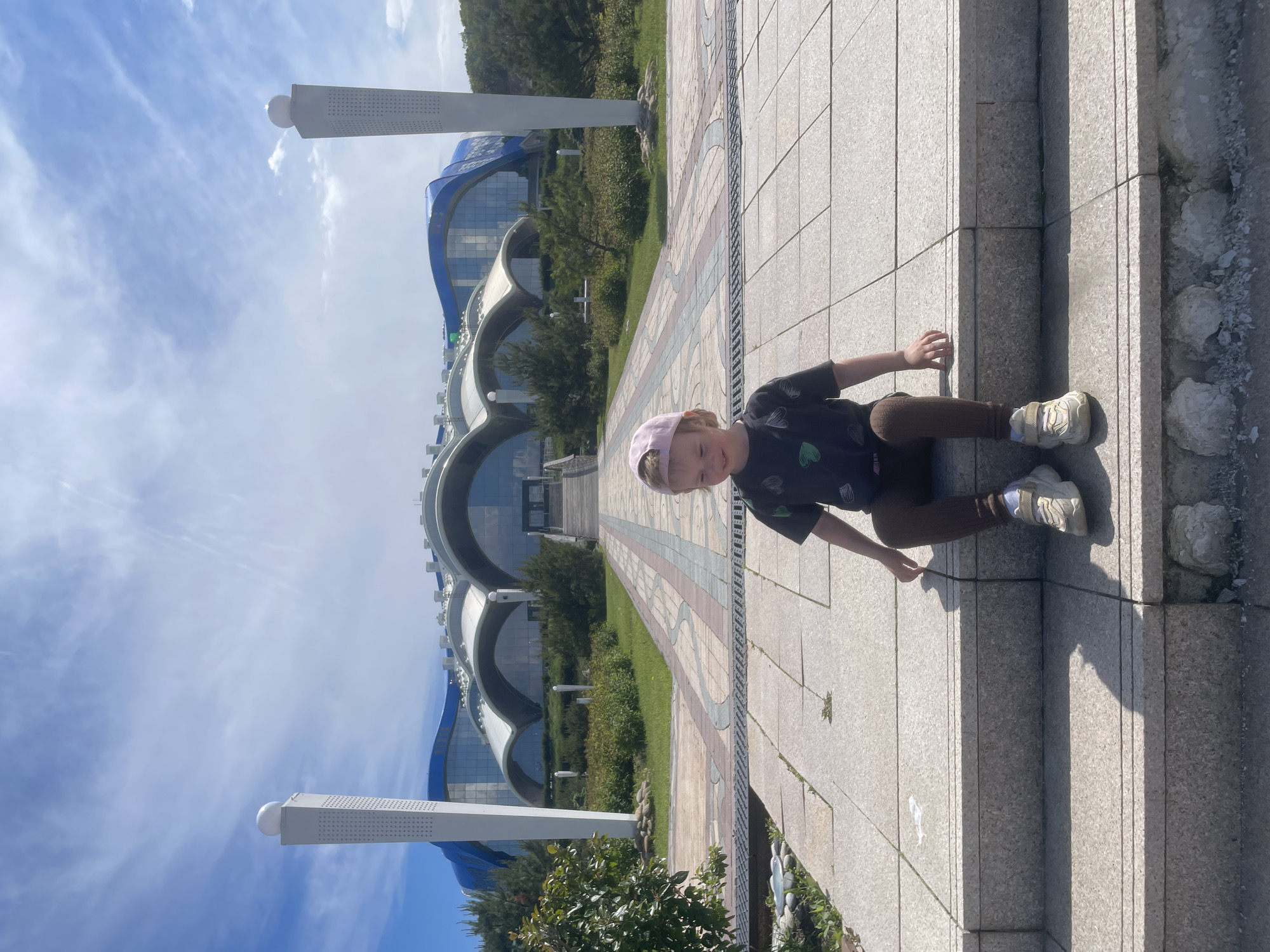 In the foreground, a young child wearing a pink baseball cap, a dark t-shirt, brown leggings, and white sneakers sits on a set of stone steps. The child is facing slightly to the left, with a subtle smile. Behind the child, a paved walkway with patterned tiles leads towards a large, modern building with a blue and white curved roof structure and glass facade. Two white towers topped with spheres flank the entrance of the building. The building appears to be the Primorye Aquarium on Russky Island, Vladivostok, Russia. The surrounding area features manicured green lawns, bushes, and trees under a bright blue sky with scattered white clouds.