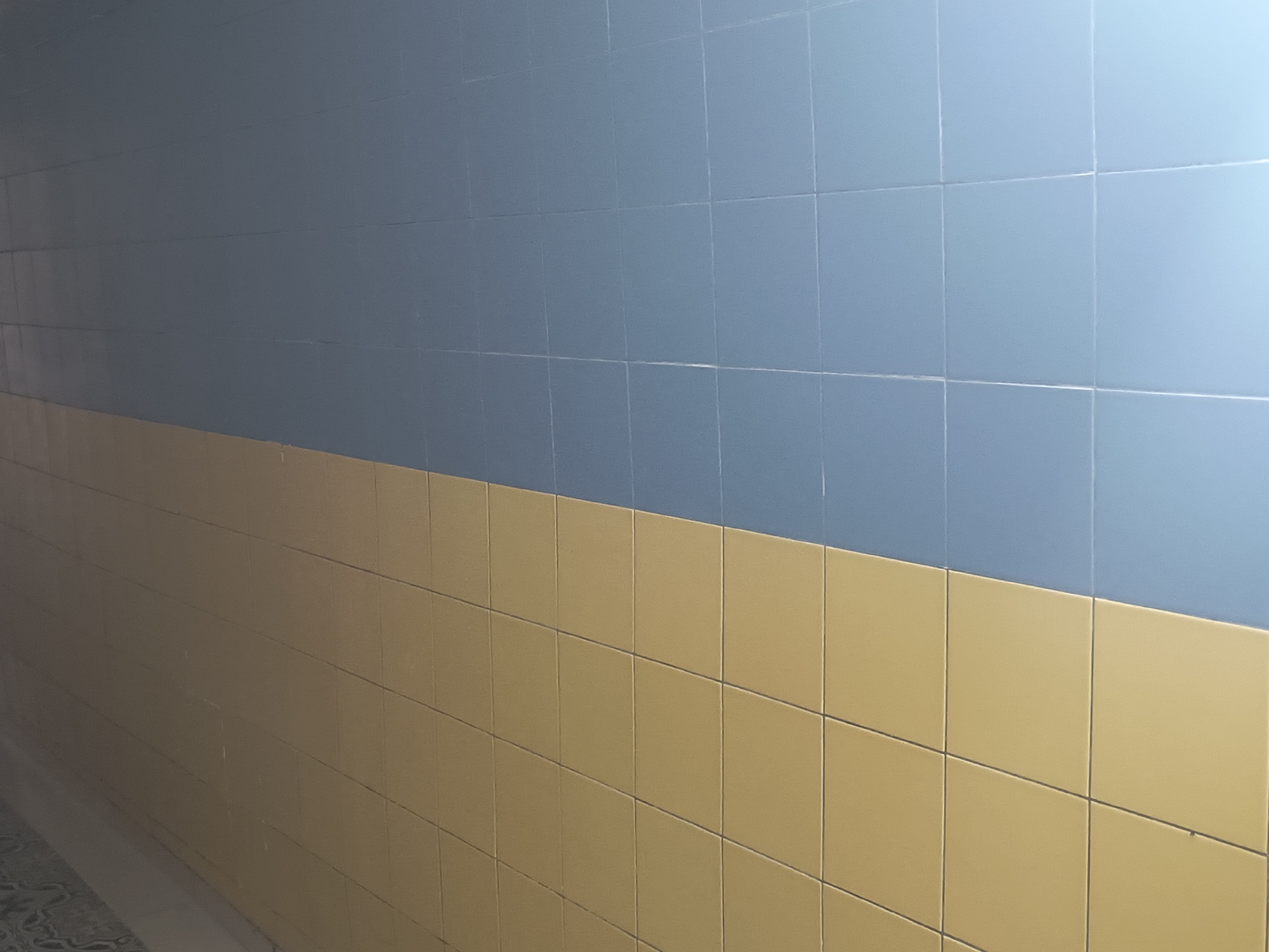 The image shows a tiled wall in what appears to be an indoor hallway or corridor. The wall is divided horizontally, with the top half tiled in a muted blue and the bottom half tiled in a light brown or tan color. The tiles are square and laid in a grid pattern with visible grout lines. The lighting suggests an artificial light source, casting subtle shadows and highlighting the texture of the tiles. The perspective indicates the viewer is standing in a corridor, looking towards a wall that recedes into the distance. There are no people or distinct objects visible in the frame other than the tiled wall itself and a hint of a patterned floor or surface at the very bottom left. The overall impression is one of a utilitarian or institutional interior space. The location in Rivne, Ukraine, is not discernible from the visual information provided in the image.