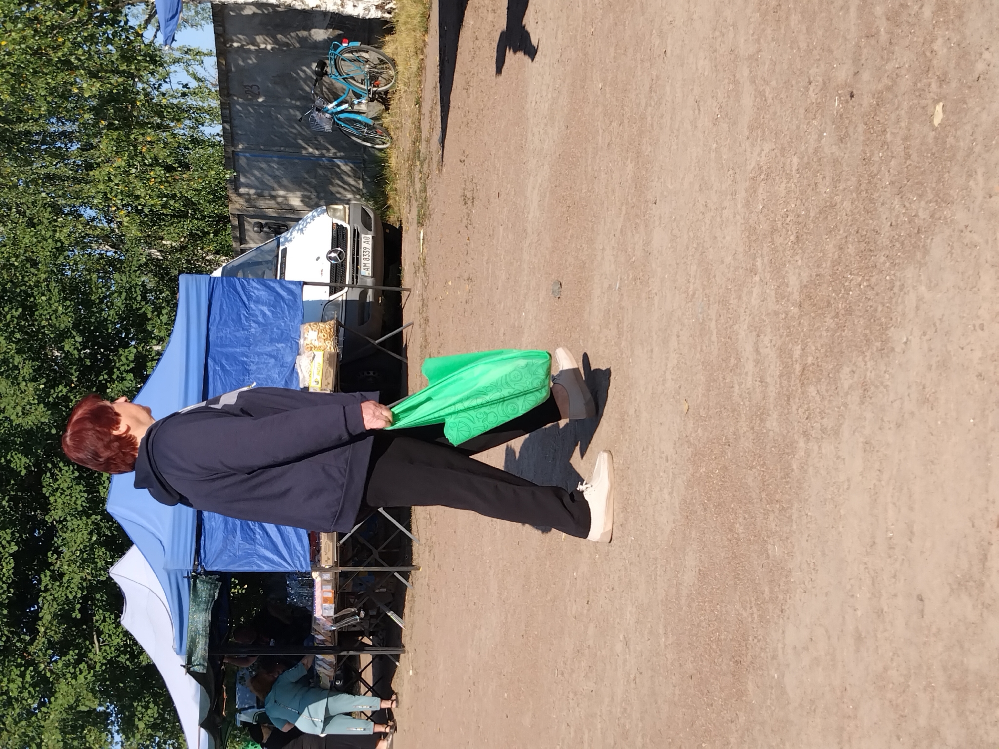 An outdoor daytime scene depicts a market in Irshansk, Ukraine. A woman with short, reddish-brown hair, wearing a dark blue hooded sweatshirt and black pants, walks from left to right, carrying a bright green reusable shopping bag. She is wearing light-colored slip-on shoes. In the background, a white van with a license plate reading "AM 8339 AO" is parked. Adjacent to the van is a light blue bicycle with a basket. Tents and market stalls are visible, with some people browsing or working. The ground is composed of dirt and gravel. Trees and a concrete wall are also present in the environment.