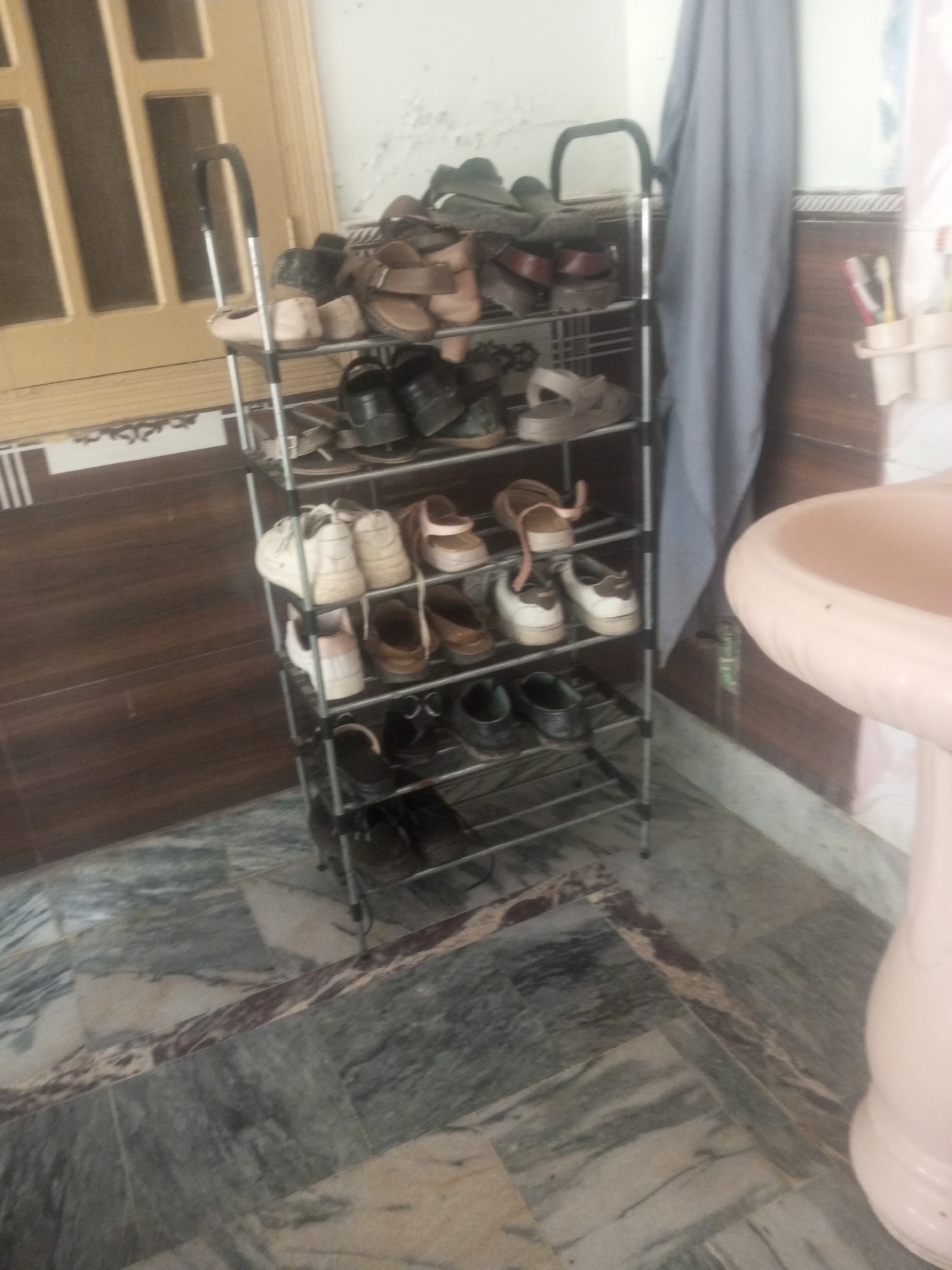 The image depicts a shoe rack with multiple pairs of footwear, suggesting an indoor setting. The shoe rack is made of metal and has several shelves filled with various types of shoes, including sandals, sneakers, and formal shoes. The flooring appears to be tiled with a marbled pattern. A pink sink is visible on the right side of the image, and a grey cloth hangs on the left. The background includes a wooden door or partition. The overall scene suggests a residential or possibly a communal living space. The presence of a shoe rack, sink, and tiled flooring are common elements in homes in Mansehra, Pakistan.