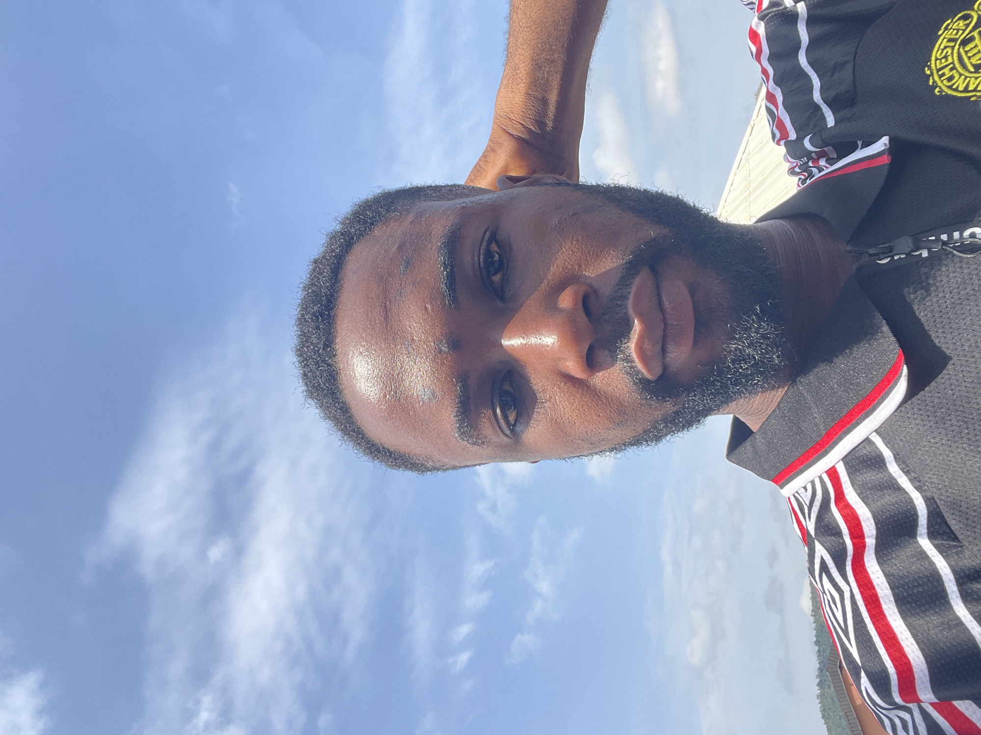 A medium shot of a man in front of a light blue sky with scattered clouds. The man has dark skin, a black beard, and black hair. He is wearing a black sports jersey with red, white, and black stripes. The jersey has the Manchester United logo on the right breast. His right hand is on his head. In the background are possible buildings. This image was taken in Lokoja, Nigeria.