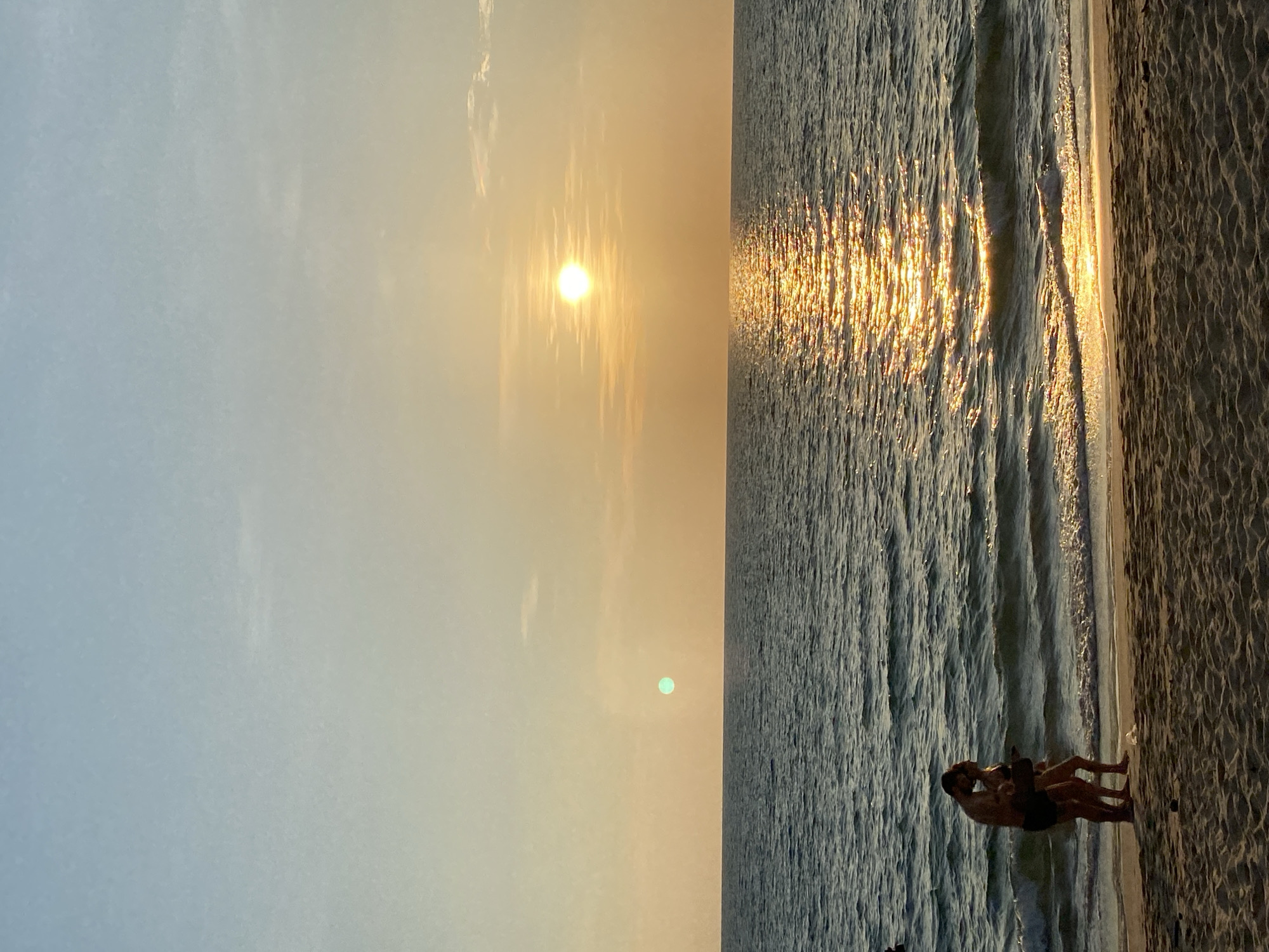 The media file captures a serene sunset beach scene, likely in Choeng Thale, Thailand, with a warm, golden hour glow. The sky, occupying the left half of the image (when correctly oriented), transitions from a pale blue on the far left to a soft orange and yellow near the center. The sun is a brilliant, bright orb positioned in the upper middle portion of the sky, casting a radiant path of light across the ocean.
The sea fills the right half of the image, with gentle waves reflecting the sun's golden light, creating a shimmering effect that stretches from the horizon towards the viewer. The texture of the water shows ripples and small waves.
On the sandy shore, located in the lower right corner where the waves meet the land, two individuals are visible. They appear to be a couple, silhouetted against the bright water and sky, standing close together in what seems to be an affectionate embrace or intimate pose, enjoying the sunset. Their figures are dark against the vivid background.
The overall atmosphere is peaceful and romantic, characteristic of an evening on a tropical beach. A faint green lens flare is noticeable in the lower part of the sky, to the left of the sun's reflection on the water. No visible text is present in the image.