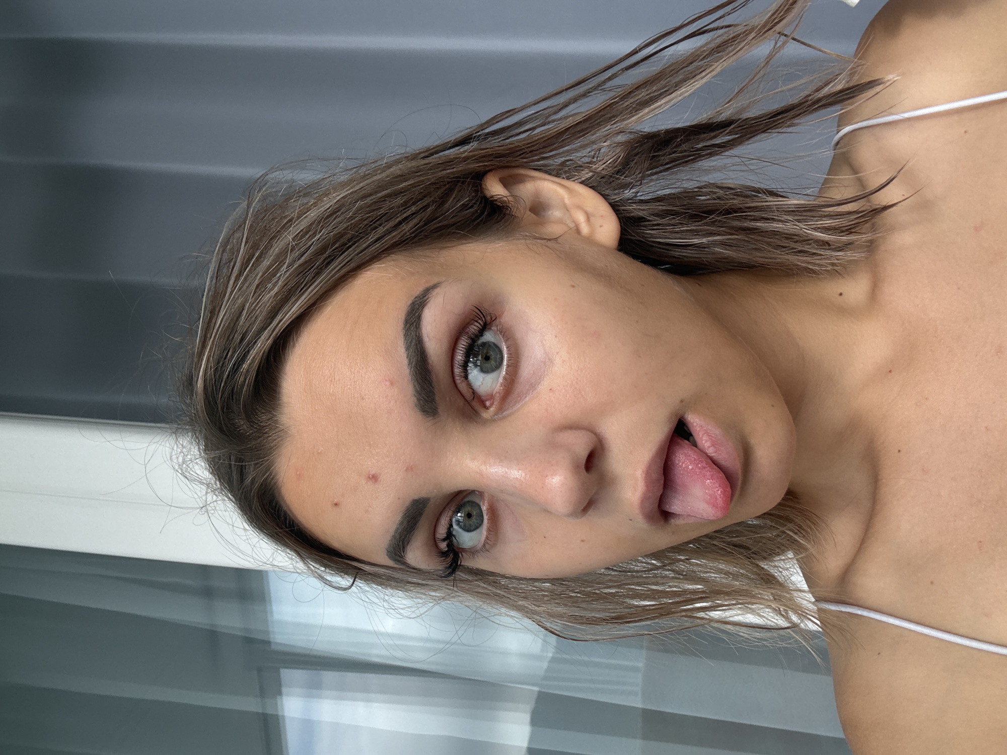 The image displays a close-up, head-and-shoulders portrait of a young woman. She has medium-length, brown hair that appears somewhat disheveled. Her eyes are open and wide, looking directly at the camera. She is sticking her tongue out, which is pink and appears slightly chapped. There are a few small blemishes visible on her forehead. She is wearing a white, spaghetti-strap top. The background consists of a light-colored, vertically striped curtain or blind on the right and a reflective surface with vertical lines, possibly a window or door, on the left. The lighting suggests it is daytime. There are no identifiable landmarks or contextual clues within the image to suggest a specific location, including Kholmsk, Russia.