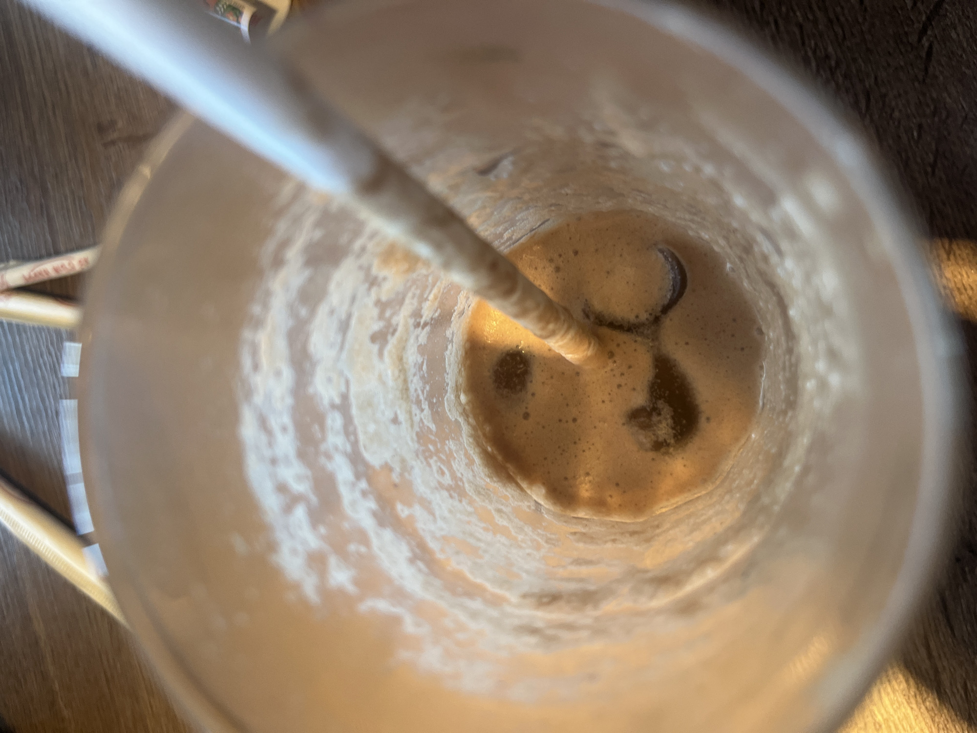 The image presents an overhead view looking down into a clear plastic cup containing a partially consumed light brown, frothy beverage, likely a coffee, chocolate milk, or smoothie. A white straw is inserted into the drink, reaching the bottom of the cup, with some of the beverage clinging to the straw itself. The liquid at the bottom of the cup shows distinct foamy bubbles and darker, possibly undissolved, sediment or chocolate chunks. The interior walls of the cup are smeared with dried residue from the drink, indicating it has been consumed for a while.
The cup rests on a wooden surface, characterized by a visible grain pattern. To the immediate left of the cup, out of focus, are several white or light-colored items, possibly additional straws or packaging. Faint red text can be discerned on one of these items, but it is too blurry to be readable; a snippet might read "FOR YOUR". The lighting suggests an indoor setting. The overall scene captures a moment of rest or completion after drinking, with the emphasis on the texture and remnants of the beverage within the cup.