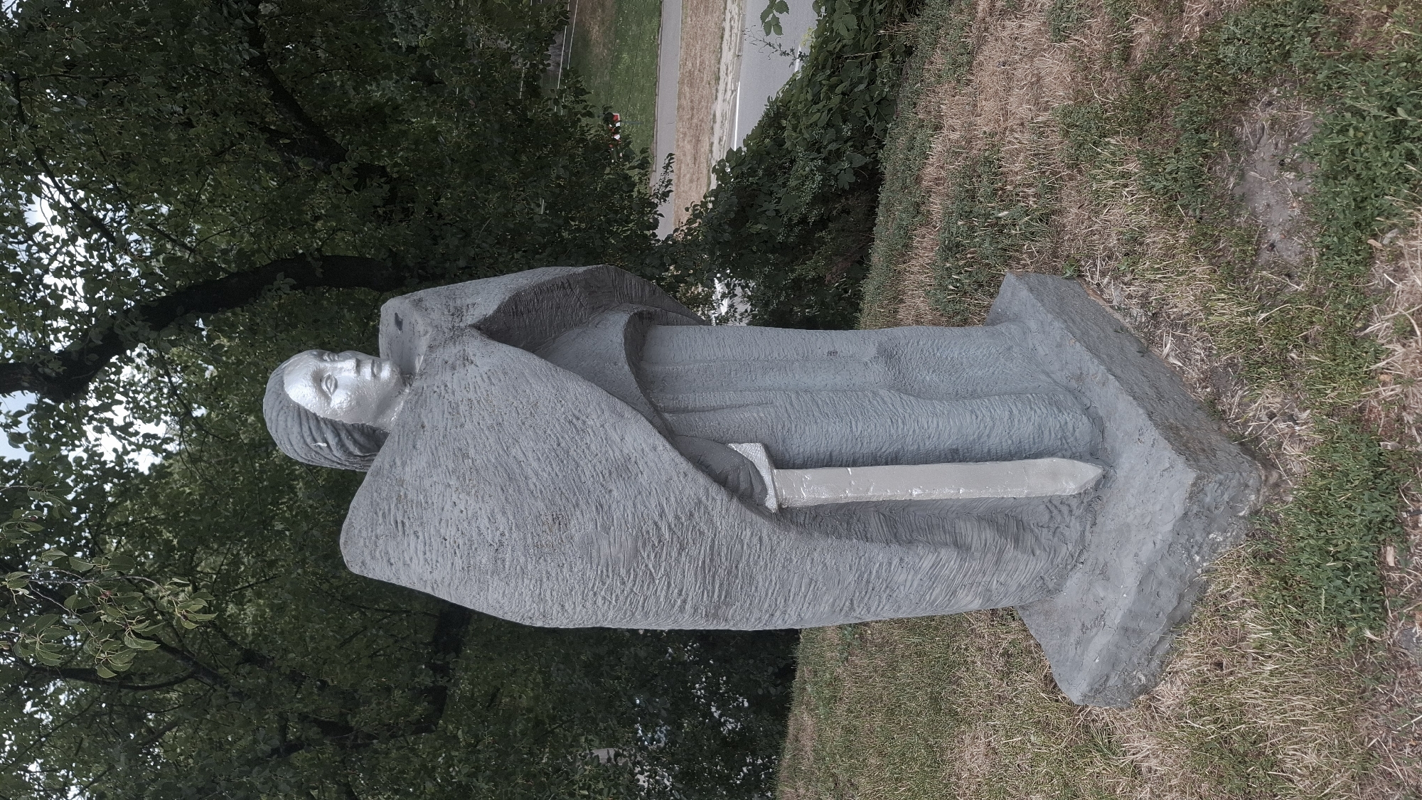 A grey, stone sculpture of a woman wearing a large cloak and holding a sword stands in a grassy area. The woman's head is tilted slightly to the right, and her facial features are visible. The sword is positioned in front of her, with the hilt extending upwards and the blade downwards. The sculpture is mounted on a rectangular stone base. In the background, there are trees with green leaves and a glimpse of a road and possibly a park area. This sculpture is located in the city of Chernihiv, Ukraine, near the historic Dytynets park.