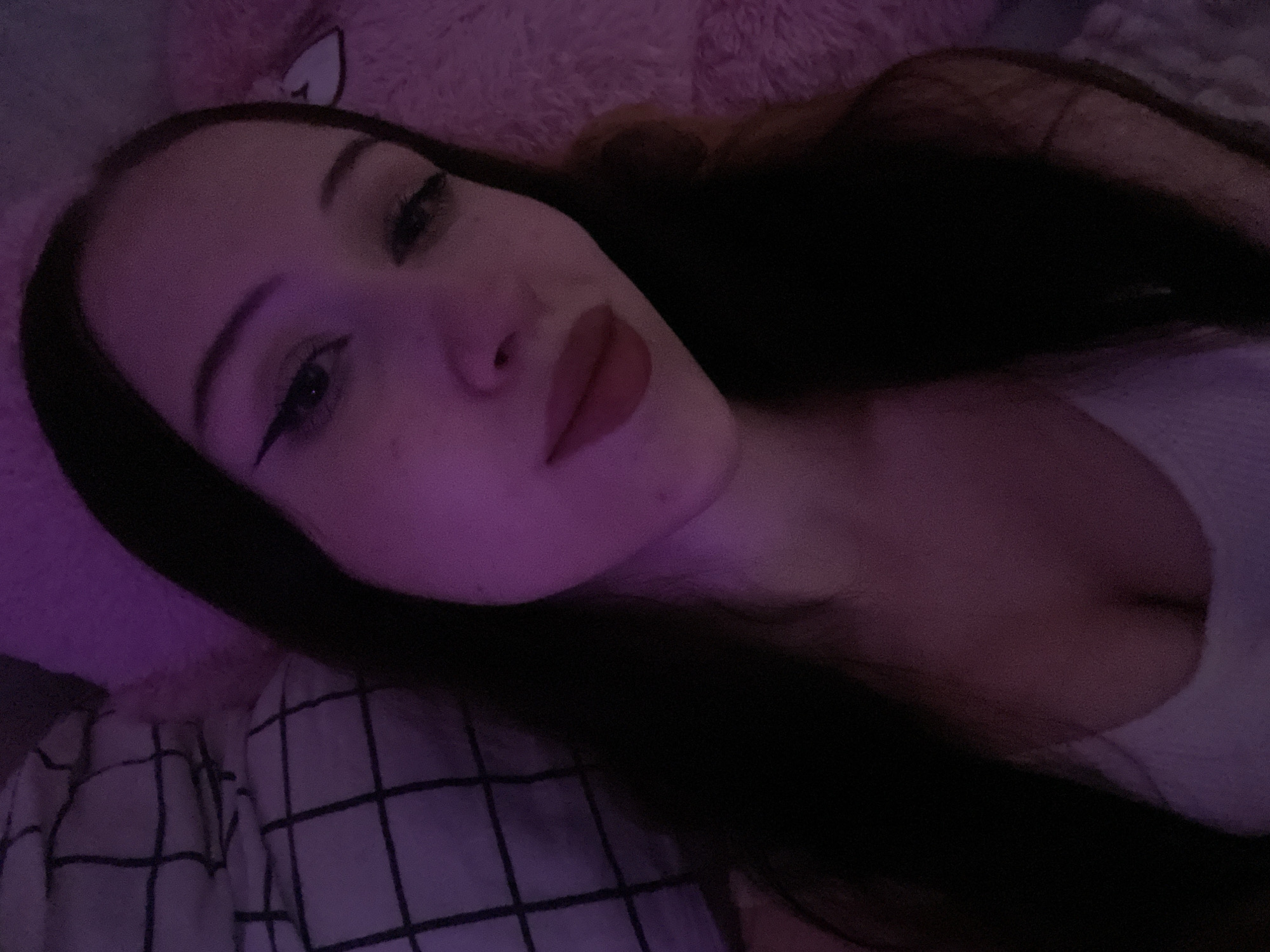 The image is a close-up, low-angle selfie of a young woman with long, dark hair and fair skin. She is wearing a white top. Her makeup includes winged eyeliner and a matte lipstick. The lighting is dim and appears to be from a purple light source, casting a pinkish-purple hue over the scene. In the background, there is a plaid patterned surface, possibly a blanket or pillow, and a fuzzy pink texture, likely another piece of bedding or a stuffed animal. The overall setting suggests a bedroom or a personal, intimate space. The image does not contain any information that would allow for a determination of its location in Votkinsk, Russia.