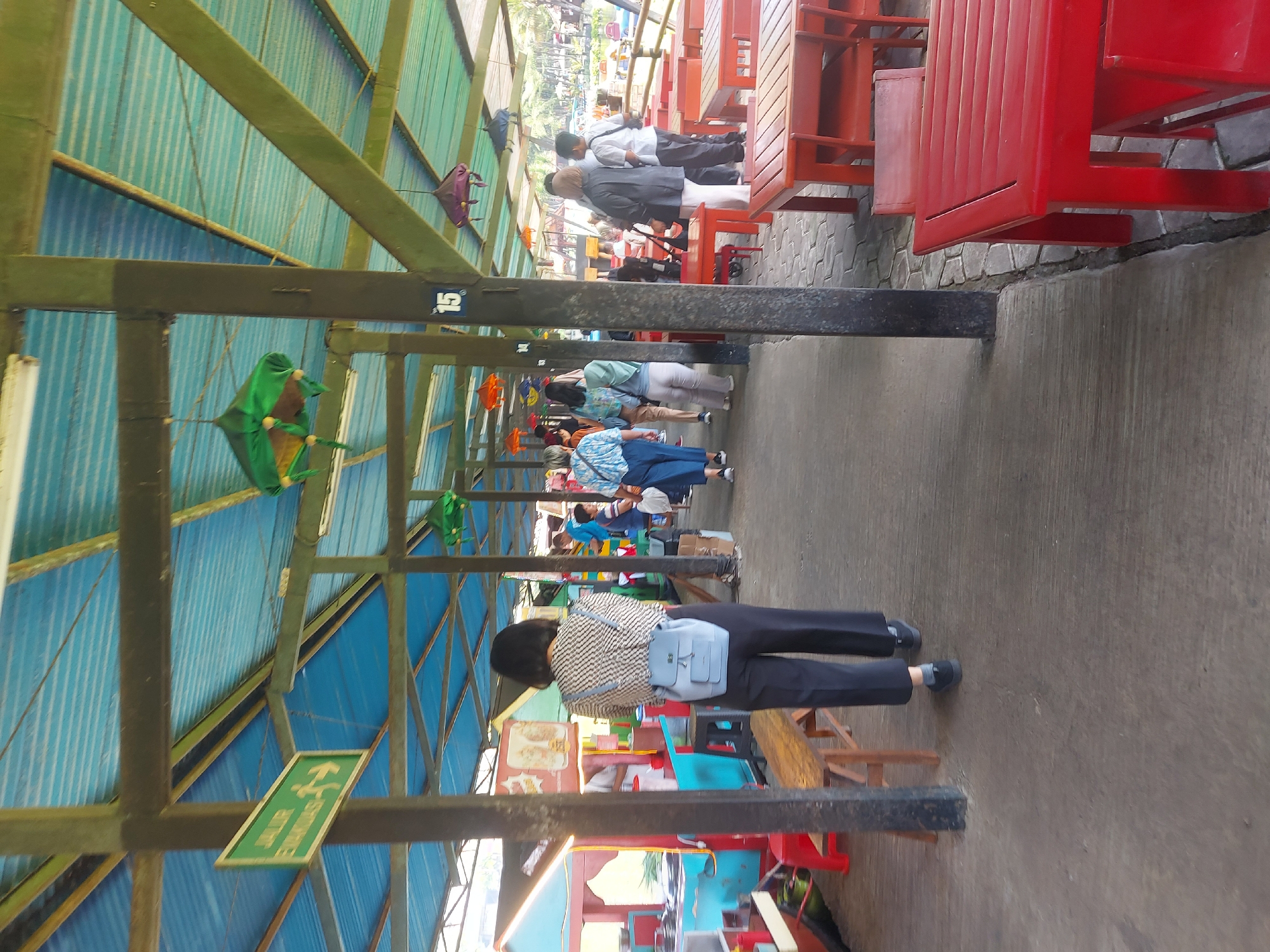 The image depicts an outdoor covered seating area at a location in Lembang, Indonesia. The structure has a corrugated blue roof supported by dark green beams. A green sign with white text and arrows is visible, indicating an exit. Hanging from the beams are various decorative items resembling upside-down umbrellas, in shades of green, orange, and purple. Several people are present, walking and standing. In the foreground, a woman from behind wearing a patterned shirt and black pants, with a light blue backpack, is walking away from the camera. Red tables and chairs are arranged in the seating area, some empty and some occupied. The ground is a mix of concrete and patterned paving stones. In the background, there appears to be a body of water and lush greenery, suggesting a scenic or recreational setting. The overall impression is a casual, open-air dining or resting area.