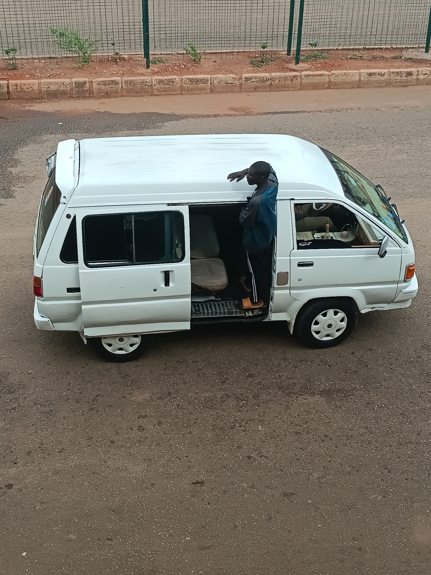An elevated shot captures a white passenger van parked on an asphalt street in Bukuru, Nigeria. A man stands in the fully open side sliding door of the van, looking towards the right side of the frame. He has dark skin and is wearing a dark blue long-sleeved top with lighter blue accents, dark trousers with white stripes, and light brown sandals. His left hand is resting on the roof of the van. The van has white hubcaps, and its interior, including worn seats, is visible through the open door and windows.
In the background, a low curb made of light-colored blocks runs parallel to the road. Behind the curb, small green plants grow in reddish-brown soil. A tall, grey metal mesh fence, supported by green poles, stands further back. The scene is illuminated by natural daylight, indicating it is daytime. No discernible text is visible. The man's position in the doorway suggests he could be a conductor, a passenger entering or exiting, or involved in public transport operations.