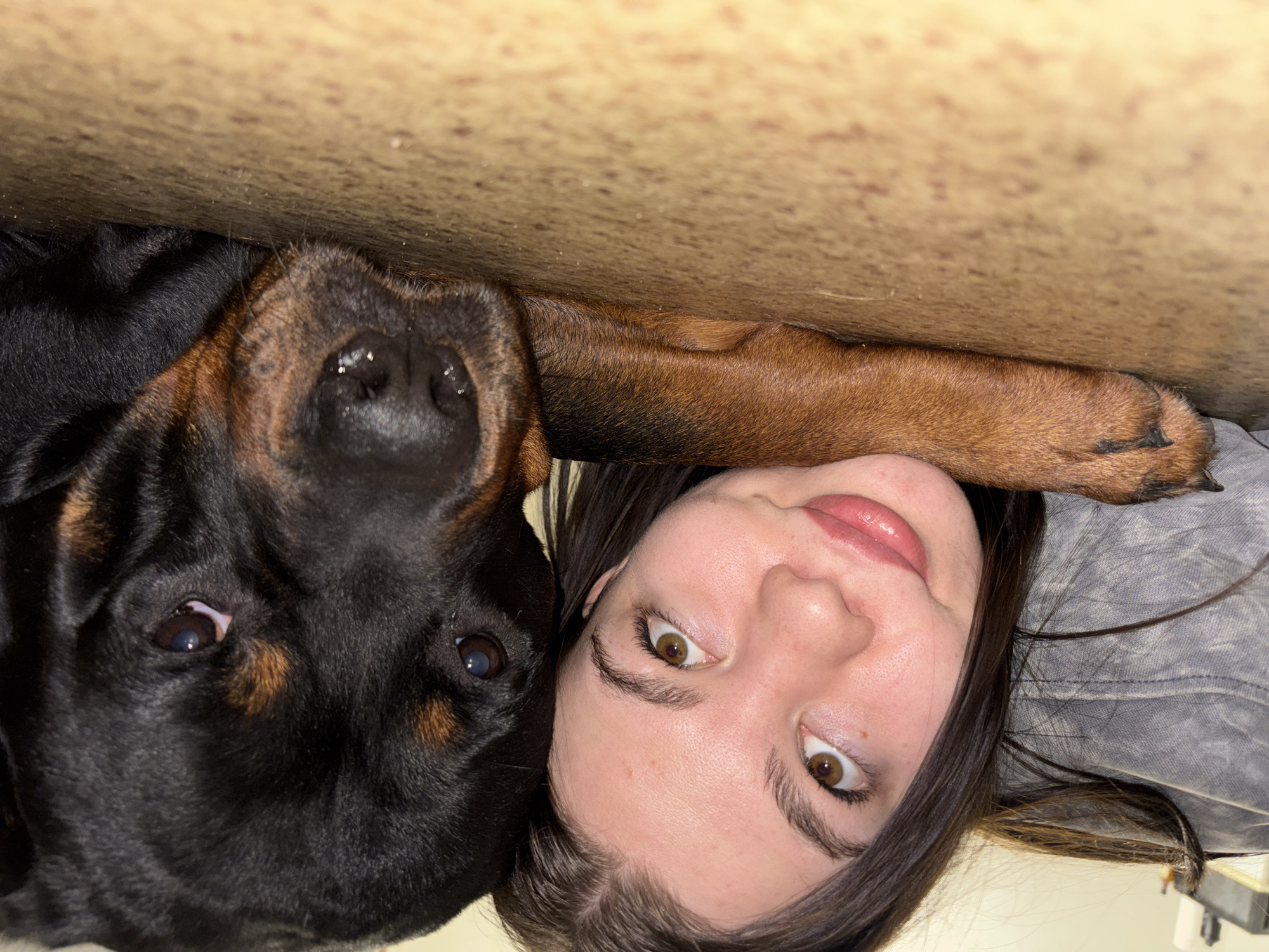 The image shows a close-up shot of a woman and a Rottweiler dog, likely taken indoors. They are positioned closely together, almost face-to-face. The woman has fair skin, brown hair, and light-colored eyes. The dog has black and brown fur, a large head, and a wet nose. Both are looking up at the camera. The background appears to be the underside of a light-colored surface. There is no evidence indicating the location is Perm, Russia.