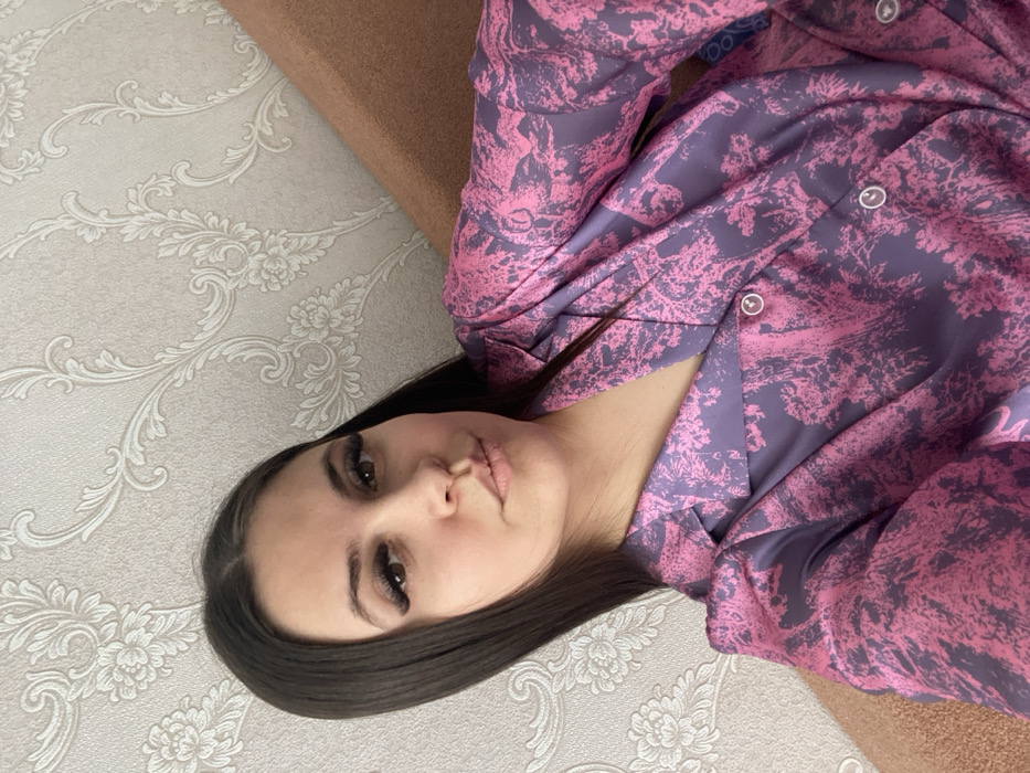 A female subject is depicted in a chest-up, frontal view, seated indoors in Chaikovsky, Russia. She has long, dark, straight hair parted centrally, with prominent eyelashes and a neutral expression, looking directly at the camera. She is wearing a collared button-up shirt with a distinctive purple and pink pattern depicting trees or foliage. White buttons are visible on the shirt's front placket. The background consists of light beige wallpaper with an ornate, scrolling floral pattern to the left, and a plain, solid brown surface, likely upholstery from a sofa or chair, to the right.