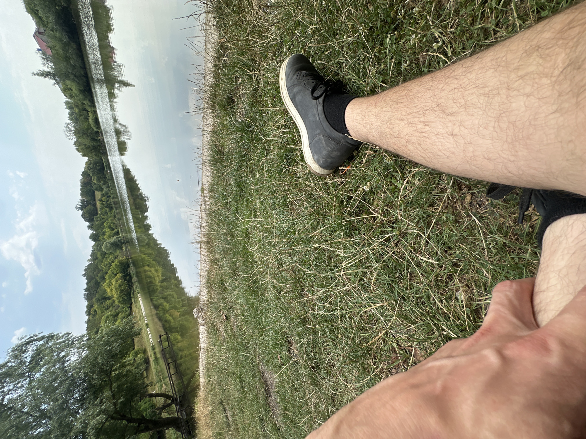 In Makiv, Ukraine, a first-person perspective shot captures a person's legs and foot resting on a grassy embankment next to a calm body of water. The person, wearing a dark shoe and black sock, is positioned with their right leg extended towards the water's edge. The background reveals a lush, tree-lined shore with reflections on the water's surface under a partly cloudy sky. A small wooden pier is visible on the left side of the image.