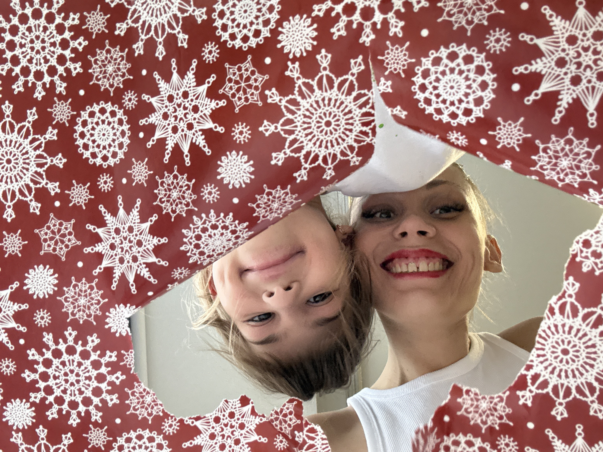 Two individuals, a child and an adult, peer down through a hole in red wrapping paper adorned with white snowflake patterns. The child's face is tilted upside down, while the adult's face is oriented upright. The adult wears a white top. The scene appears to be indoors. No information is available to confirm the location is in Fedorino, Russia.
