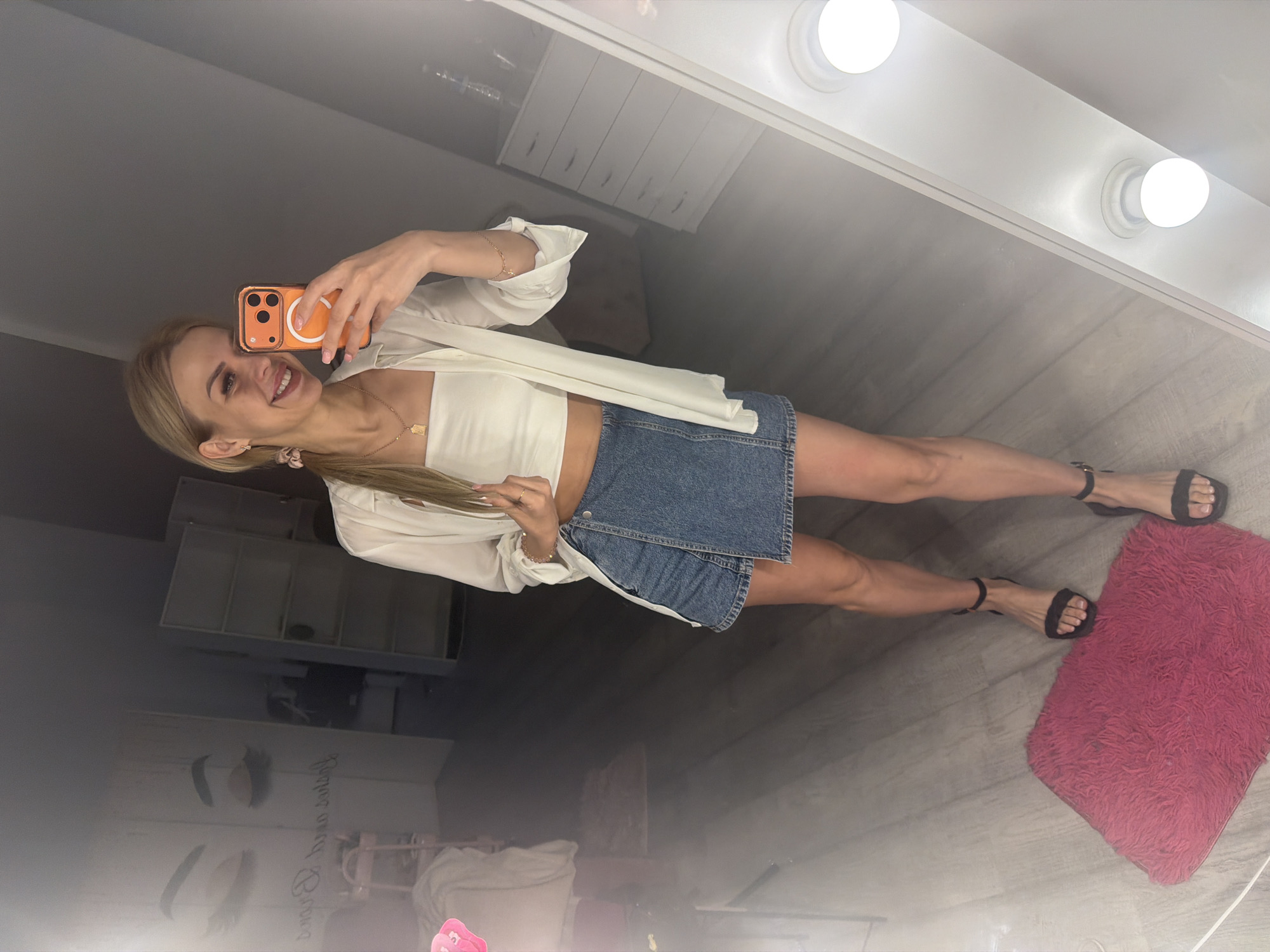 A blonde woman, wearing a white crop top, an unbuttoned white shirt, and a denim skort, stands in front of a mirror, taking a selfie with her orange-cased iPhone. She has on black high-heeled sandals and a gold necklace. She is indoors, in what appears to be a room with wooden floors, a pink rug, and a large mirror with lights. The background includes a dresser, shelves, and a poster of eyelashes. The overall atmosphere suggests a dressing room or a beauty salon. The location is identified as Kyiv, Ukraine.