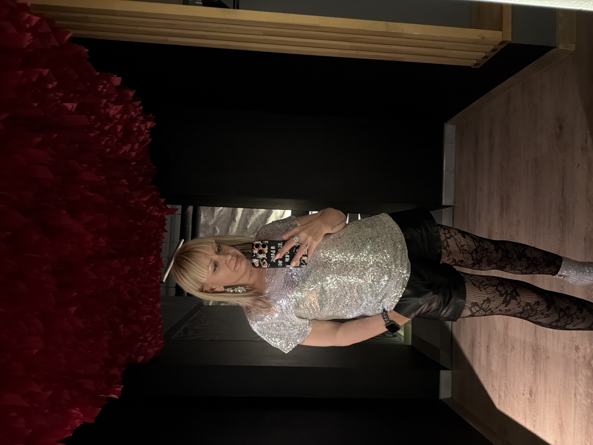 A woman is standing in what appears to be a hallway or corridor. She is wearing a silver sequined top, black leather shorts, patterned black tights, and silver boots. She is holding a phone and taking a selfie. A wall covered in red, textured material is visible on the left side of the image. The hallway has wooden floors. This image cannot be definitively traced to Chaikovsky, Russia based on visual evidence.