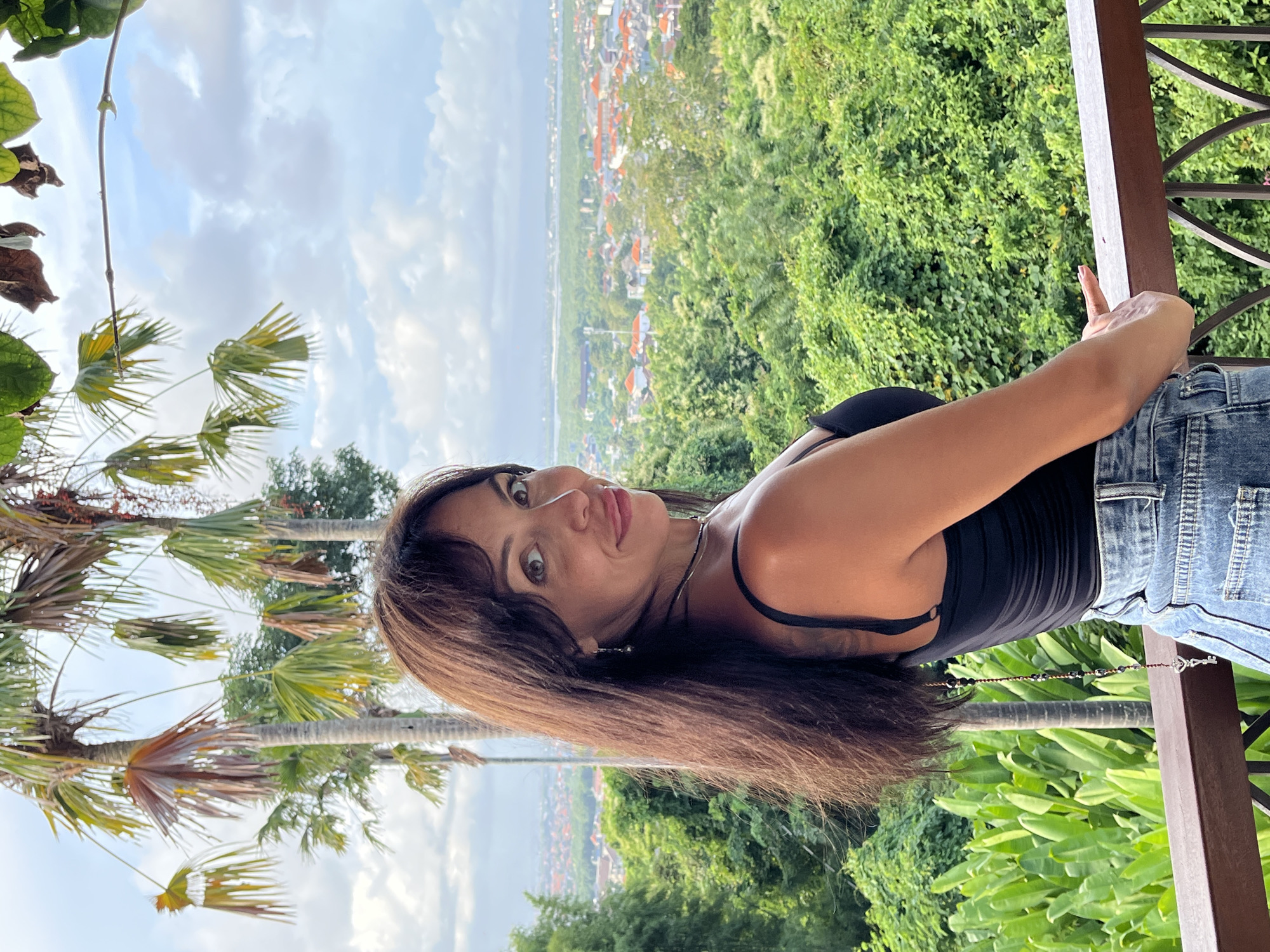 A brunette woman stands on a balcony, seemingly overlooking Kuta Selatan, Indonesia. She is wearing a black tank top and denim jeans. She is leaning against the wooden railing of the balcony. Behind her, there is a dense forest, and in the distance, buildings that appear to be part of the city can be seen. There are also palm trees visible to the left. The sky is partly cloudy.