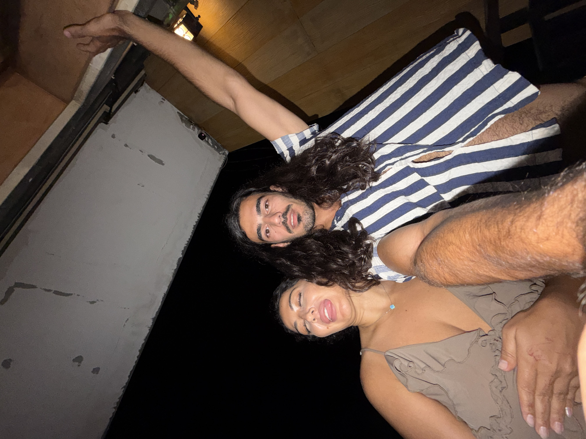 This close-up image, likely a selfie, captures two individuals, a man and a woman, posing playfully in North Kuta, Indonesia, during what appears to be nighttime. The photo is tilted, suggesting a dynamic, low-angle shot.
The man, positioned slightly behind and above the woman, has long, dark, curly hair and a beard. He is wearing a blue and white striped shirt and looks directly at the camera with a subtle smile. His right arm is extended upwards and bent, with his hand reaching towards the upper frame.
In the foreground, the woman has long, dark, curly hair and is wearing a light brown, ruffled top. She has a delicate necklace with a light blue pendant. She is making a humorous expression, sticking out her tongue and partially closing her eyes. A hand, likely the man's, is visible resting near her.
The background is dimly lit, with a decorative lantern fixture visible in the upper right, indicating the use of artificial light. Elements of the setting include a wooden ceiling or structure on the right and a grey, textured wall with some marks on the left, fading into darkness. The overall mood is casual, playful, and affectionate. There is no visible text in the image.