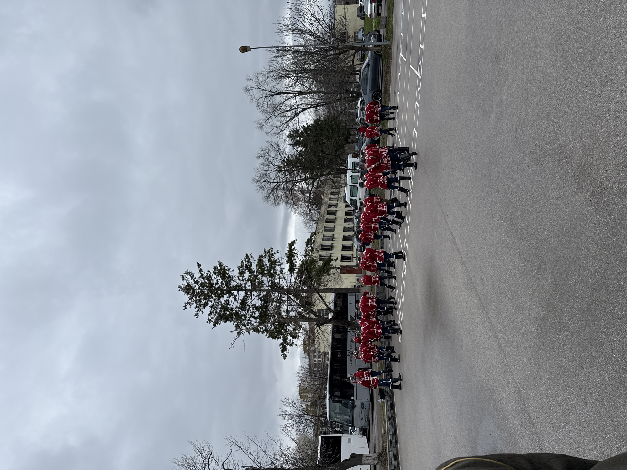 A line of individuals in red jackets and dark pants with tall dark hats are marching in a parking lot. The parking lot has white painted lines and is adjacent to a row of parked vehicles. A building is visible in the background. The weather is overcast. A bus is parked near the marching individuals. The building in the background suggests an urban location, potentially in Sofia, Bulgaria.