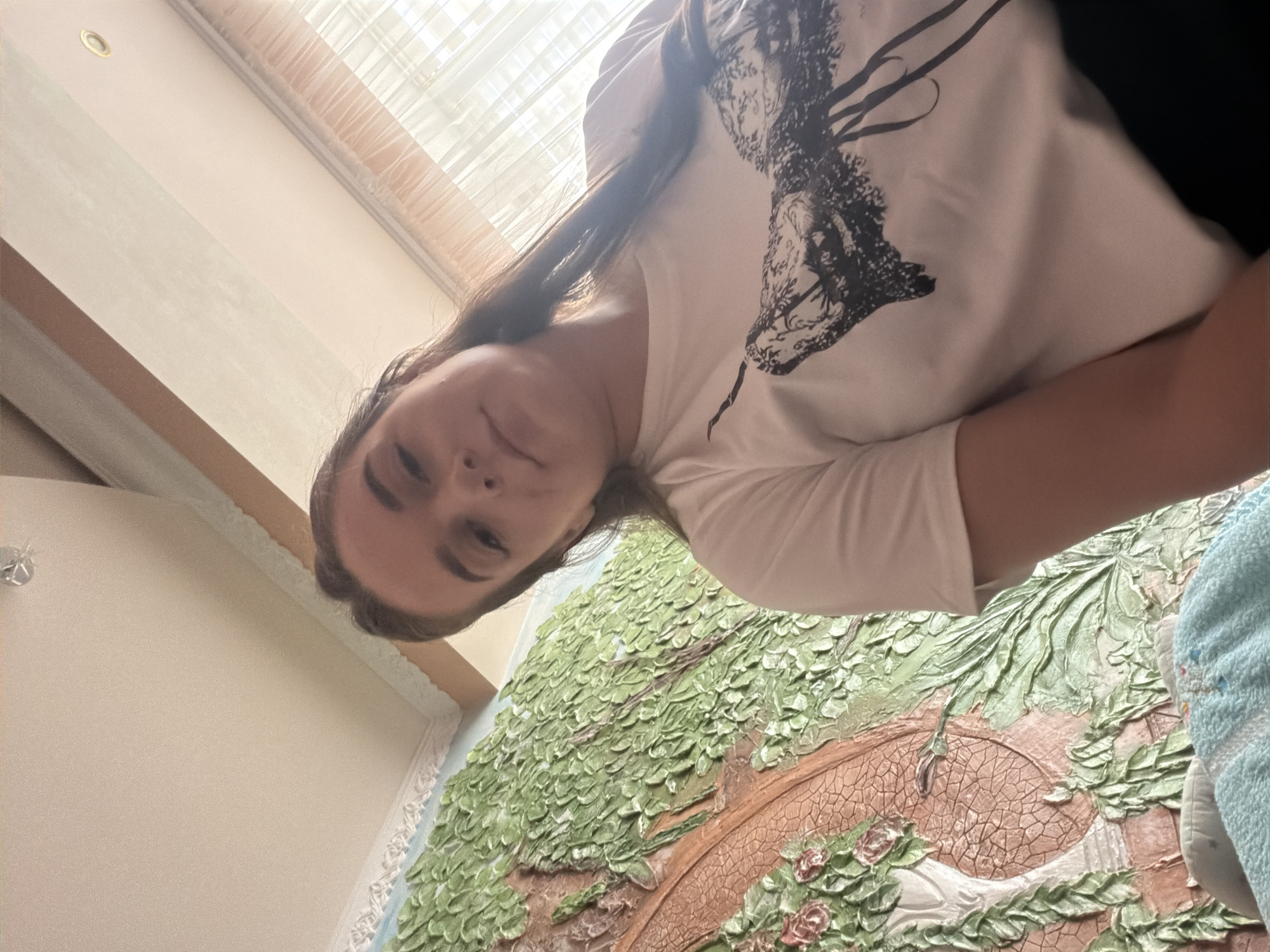 A young woman with long, dark brown hair is shown from the chest up, looking directly at the camera. She is wearing a white t-shirt with a black graphic design. To her left is a textured wall decoration depicting a garden scene with green foliage, a pathway, and pink roses. The environment appears to be indoors, with a ceiling visible above, featuring light fixtures. The overall lighting suggests it is daytime, with natural light filtering in from a window to the right. The location is identified as Durlești, Moldova.