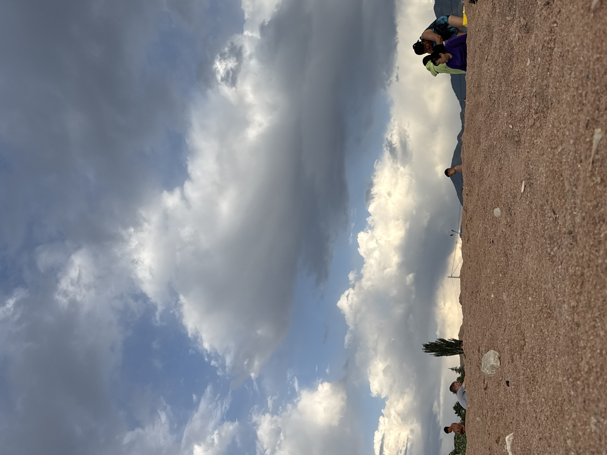 In Каджи-Сай 2, Kyrgyzstan, an outdoor scene unfolds with a sandy foreground and a dramatic sky. Several people are present on a sandy hill. In the mid-ground, a child in a grey shirt is leaning forward. To the left, a child wearing a dark shirt stands near the edge of the hill. Further to the left, another child is visible. On the right side of the mid-ground, a group of three people are interacting. One person in a blue swimsuit appears to be interacting with another person in a purple top, who is partially obscured by a person in a green garment. In the background, mountains are visible, and a tall, slender tree stands prominently. The sky is filled with a mix of dark, ominous storm clouds and brighter, illuminated clouds, with patches of blue sky peeking through. The sand in the foreground is a reddish-brown color and appears to be coarse.