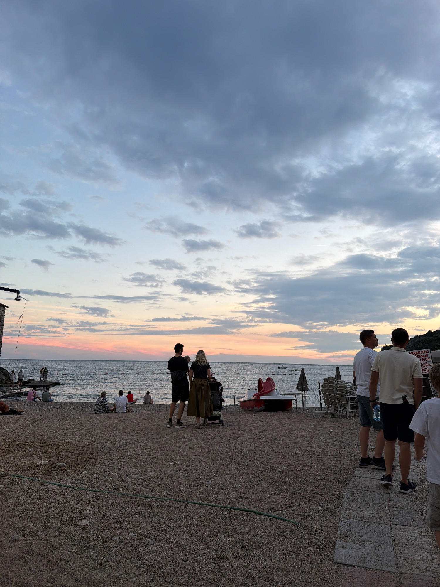 The image depicts a sunset scene on a pebble beach in Budva, Montenegro. The sky is filled with dramatic clouds, transitioning from pale blue at the top to soft pink and orange hues near the horizon. The sea is calm with a few distant boats visible.
In the foreground, several people are scattered on the beach. On the right side, three men stand together, facing away from the camera and towards the sea. One man on the far right is partially visible and appears to be a child. To their left, a couple stands, the man facing the sea and the woman looking towards the man. A baby stroller is positioned near them. Further to the left, on the water's edge, a group of people are seated on rocks and the shore. On the far left, a building structure with a hanging pipe and flag is visible. The beach itself is made of small pebbles and sand, with some scattered rocks. A green hose lies on the ground in the middle of the frame. In the middle ground, a red paddle boat and some beach chairs are visible. The overall atmosphere is one of a relaxed evening at the coast.
