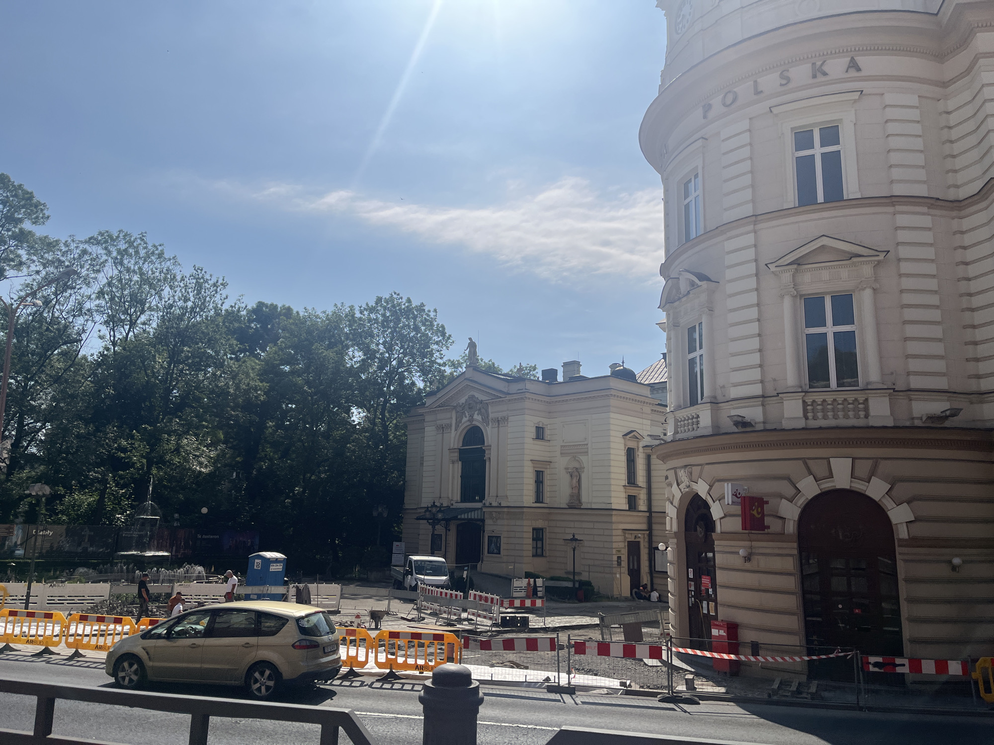 The image shows a street scene in a city with historical architecture and ongoing construction. In the foreground, a light-colored minivan is parked on the side of the road, partially obscured by orange safety barriers. The barriers, some with red and white markings, indicate a construction zone.
Behind the barriers, a yellow-colored minivan is driving on the street. Several people are visible in the background, some standing near the minivan, others near a portable toilet unit. A building with a classical facade, featuring arched doorways and windows, stands on the right side of the image. Above one of its rounded sections, the word "POLSKA" is inscribed.
To the left of this building, a lighter-colored, rectangular building with a prominent entrance and arched windows is visible. Further in the background, dense green trees line a park area, where a fountain is also discernible. The sky is blue with scattered clouds, and the sun is shining brightly, creating some lens flare in the upper part of the image. The overall impression is a sunny day in a city square, with public works underway.