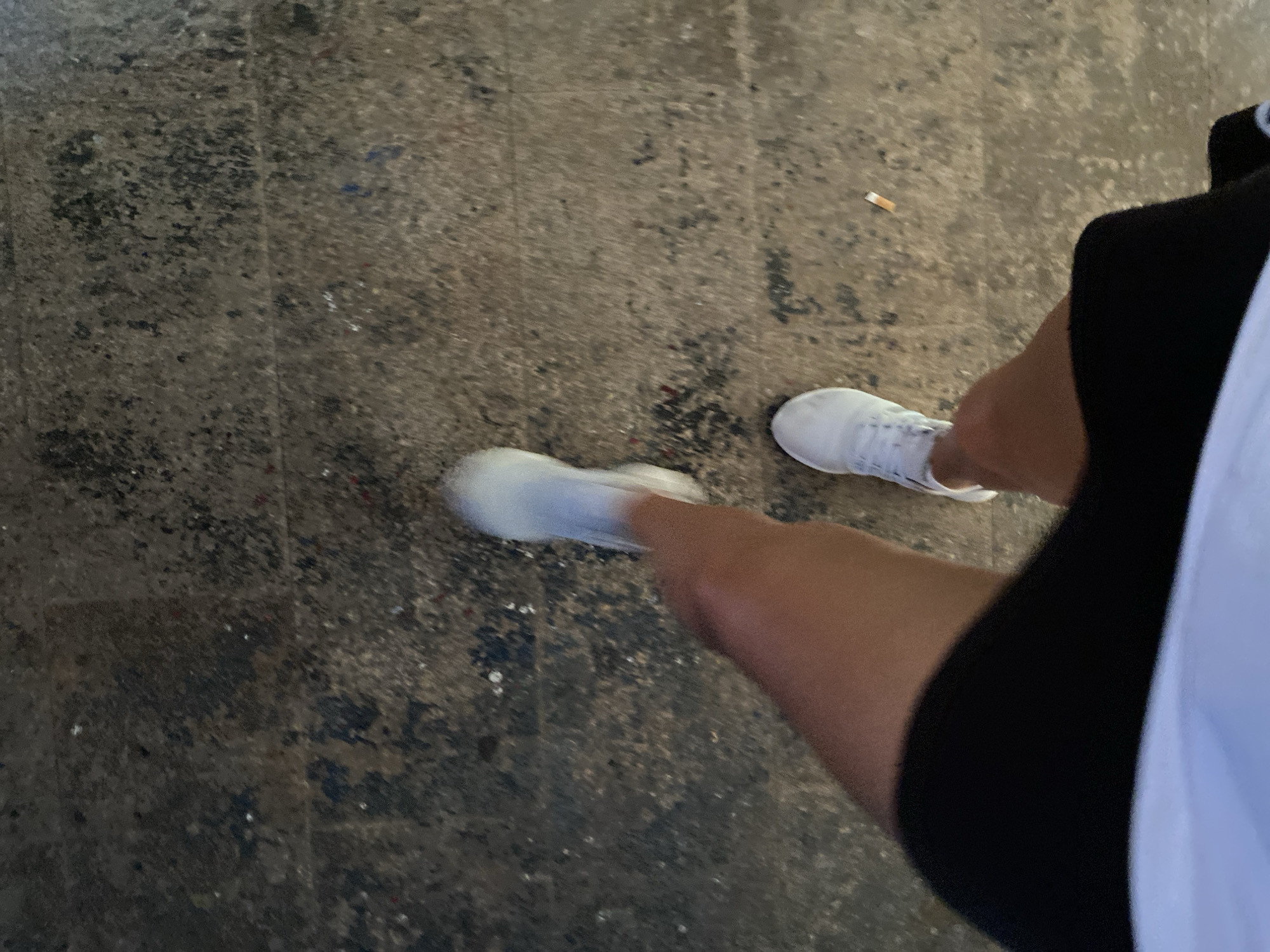 The image shows a low-angle, first-person perspective of a person's legs and feet walking on a worn, speckled concrete surface. The ground appears to be a sidewalk or pavement, with visible cracks and discolorations suggesting age and weathering. There are scattered small particles of debris on the ground. The person is wearing white athletic shoes and black shorts. The left leg is in motion, indicated by slight blurring. In the mid-right portion of the image, a discarded cigarette butt is visible on the ground. There is no specific visual information in the image to suggest the location within the city of Aksu, Turkey.