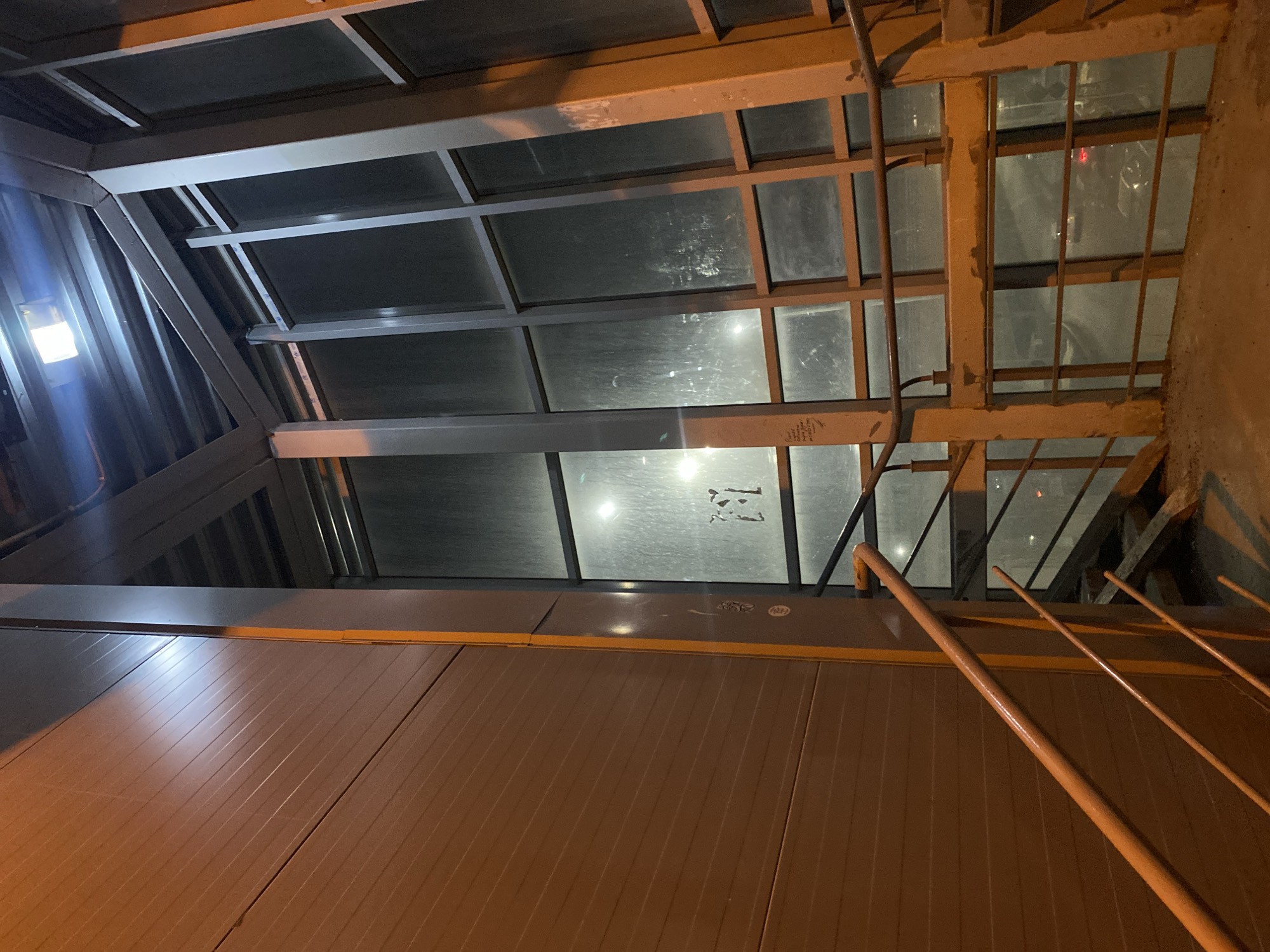 The image depicts an elevated pedestrian walkway with a metal and glass enclosure. The walkway appears to be constructed with a steel framework, supporting panes of glass that form the roof and sides. The metal framework is painted in a light color, possibly a beige or light gray. The glass panes are somewhat obscured, suggesting they may be frosted or dirty.
Below the walkway, a wall with horizontal ribbing is visible, suggesting a pedestrian tunnel.
The presence of the glass enclosure and the ribbed tunnel, suggests a location near an underpass in the city of Kazan, Russia.