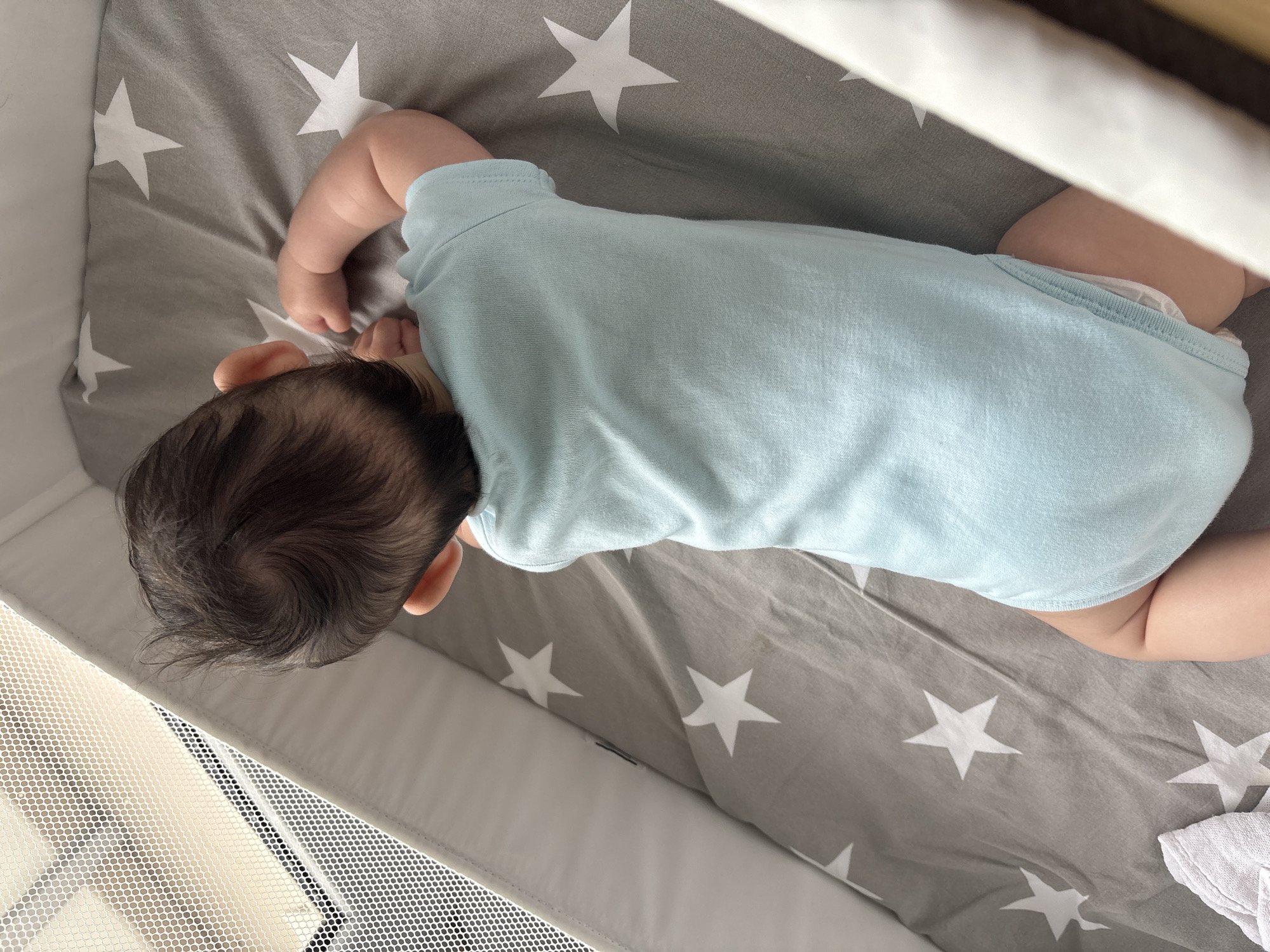 An infant, viewed from above, is lying on their stomach in a crib. The infant is wearing a light blue onesie and a diaper. The crib bedding is gray with white stars. The crib has a white mesh netting on one side. The scene takes place indoors, likely in a residential setting, and no specific location in Plovdiv, Bulgaria, can be determined from the image.